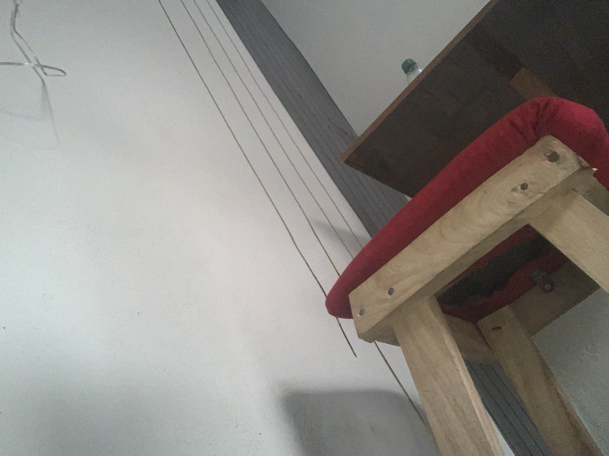 The image depicts a close-up view of a wooden chair with a red cushion, positioned against a textured white wall. The chair appears to be partially disassembled or in a state of disrepair, with visible wooden beams and screws. Several thin, light-colored wires are suspended horizontally across the upper portion of the image, running parallel to a grey horizontal band that separates the wall from a darker surface, possibly a ceiling or a raised platform. The lighting suggests an indoor environment. There is no discernible information in the image to ascertain its location within the city of Jauro Yinu, Nigeria, nor are there any people or specific actions depicted.