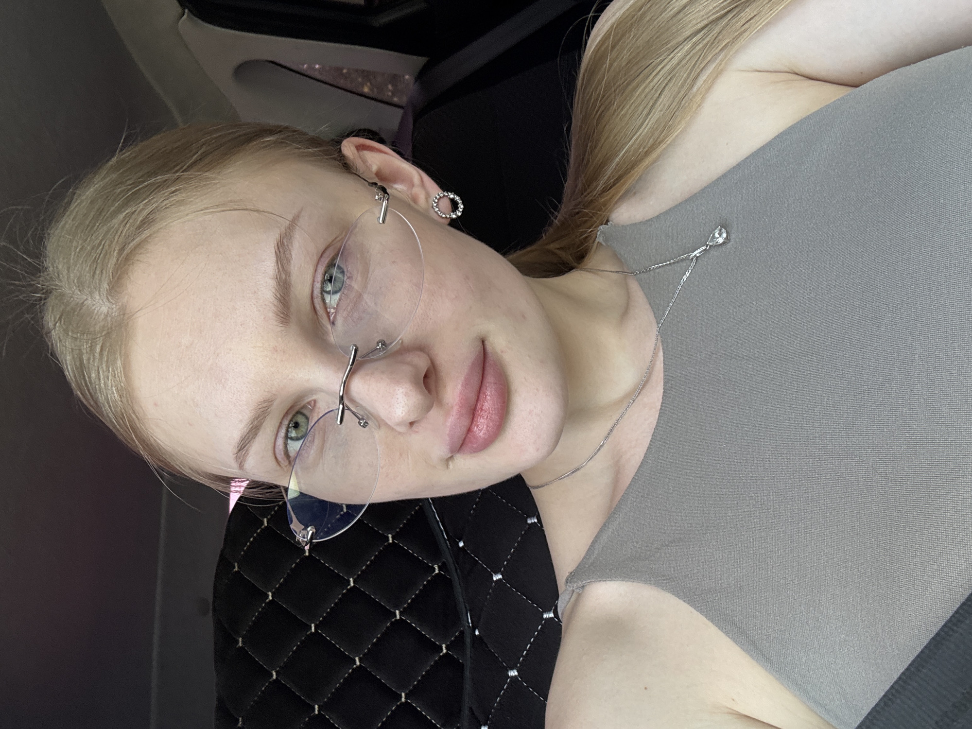 A fair-skinned, blond woman with light eyes wearing frameless glasses, a silver necklace, and a gray top is pictured in a close-up selfie. Her hair is pulled back in a loose ponytail. The background appears to be the interior of a car, with a quilted, dark material visible behind her. The location is likely Perm, Russia as indicated by the user's prompt.