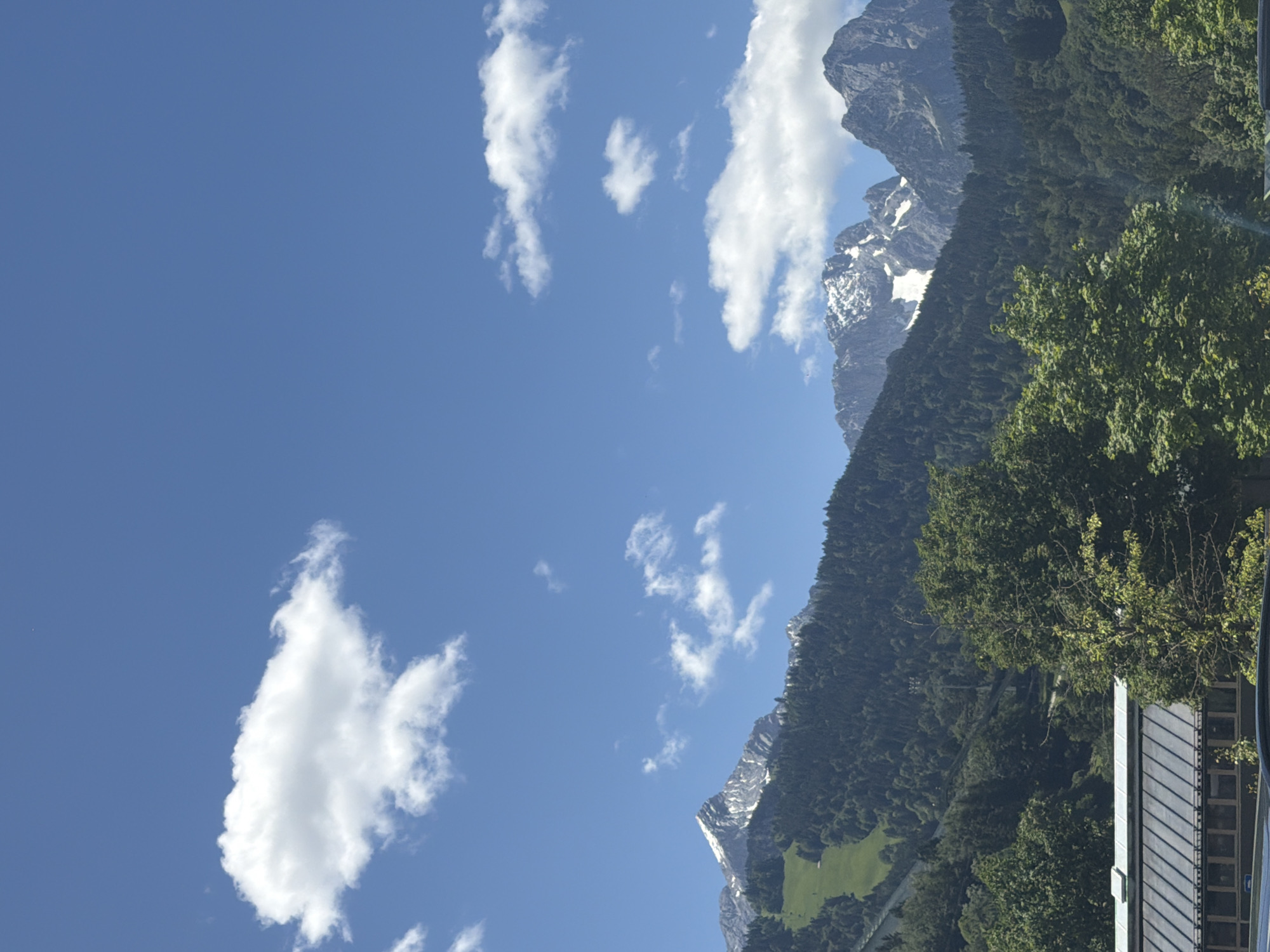 The image shows a mountain range on a clear day. The sky is a bright blue with scattered white clouds. The mountains are covered in dense green forests, with some snow-capped peaks visible at the top. In the foreground, there is a building with a sloped roof. The scene appears to be a landscape photograph taken from a distance, capturing the grandeur of the mountains.