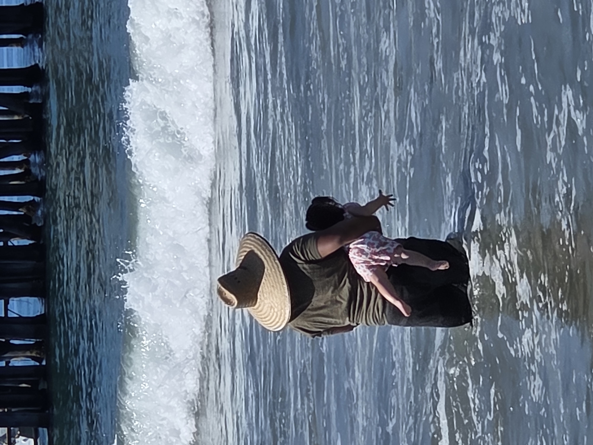 A person wearing a wide-brimmed straw hat and a green shirt stands in the ocean, holding a baby in their arms. The baby is wearing a pink and white patterned dress and is reaching out. Waves are breaking in the background. In the distance, pilings of a pier are visible. The scene is likely Santa Monica, California, due to the presence of the pier.