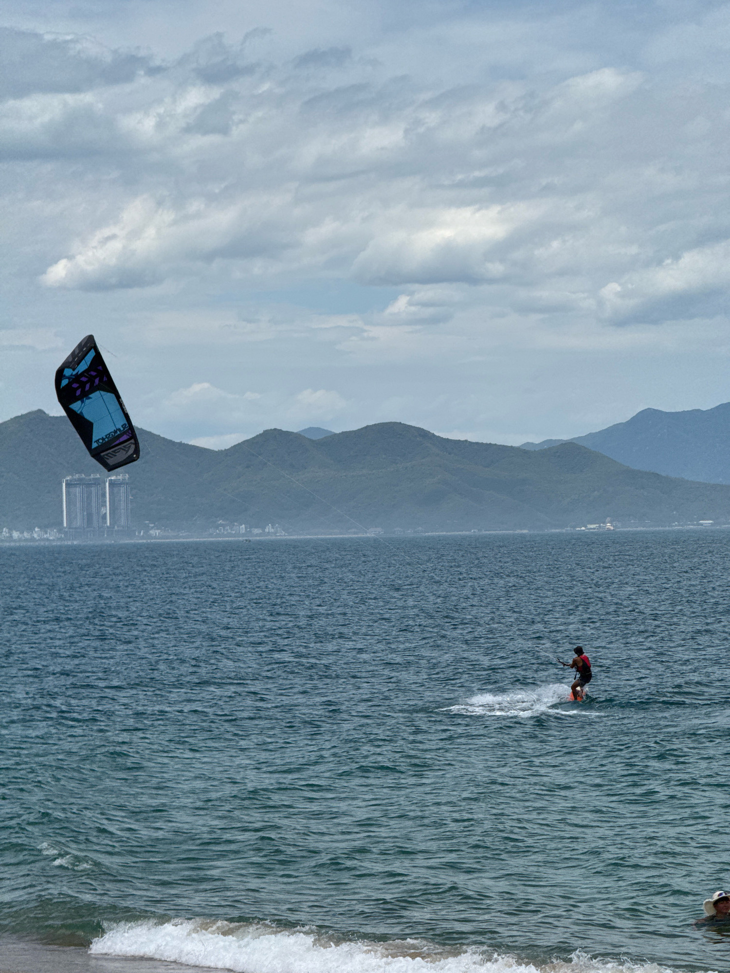 In the city of Nha Trang, Vietnam, under a cloudy sky, a person is kitesurfing on the ocean. The kitesurfer, wearing a red life vest and dark shorts, is positioned to the right of the frame, standing on a board and being pulled by a large, blue and black kite visible in the upper left quadrant. The kite has purple geometric patterns and appears to be branded. The water is a deep blue with moderate waves. In the background, a line of green, mountainous terrain stretches across the horizon. Visible on the land are several tall, modern buildings, suggesting an urban coastline. The foreground shows the edge of a sandy beach with a small wave breaking.