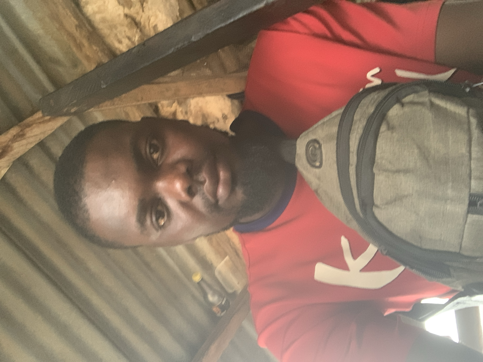 A close-up, waist-up selfie of a Black man is captured, looking directly at the camera. He is wearing a red t-shirt with a blue trim around the collar and what appears to be a white graphic on the left chest. A gray sling bag is worn across his chest, with its zipper partially visible. The man has short, dark hair and a beard.
The background suggests an indoor, rustic environment. The ceiling is made of corrugated metal sheeting supported by wooden beams. To the right, there is a rough, stone or brick wall. A dark wooden beam angles downwards from the upper right to the middle right of the frame, partially obscuring the wall. A bottle with a yellow cap and some other objects are visible on a shelf or surface in the upper left background, behind the man's left shoulder.
The lighting is somewhat dim, casting a slight haze over the image. The overall impression is casual and personal, consistent with a selfie taken in a home or informal setting. The presence of corrugated metal roofing and rough walls could be indicative of a dwelling or workshop in an area with less developed infrastructure. Given the reference to Shagamu, Nigeria, such an environment is plausible, potentially within a residential compound or a local business premises.