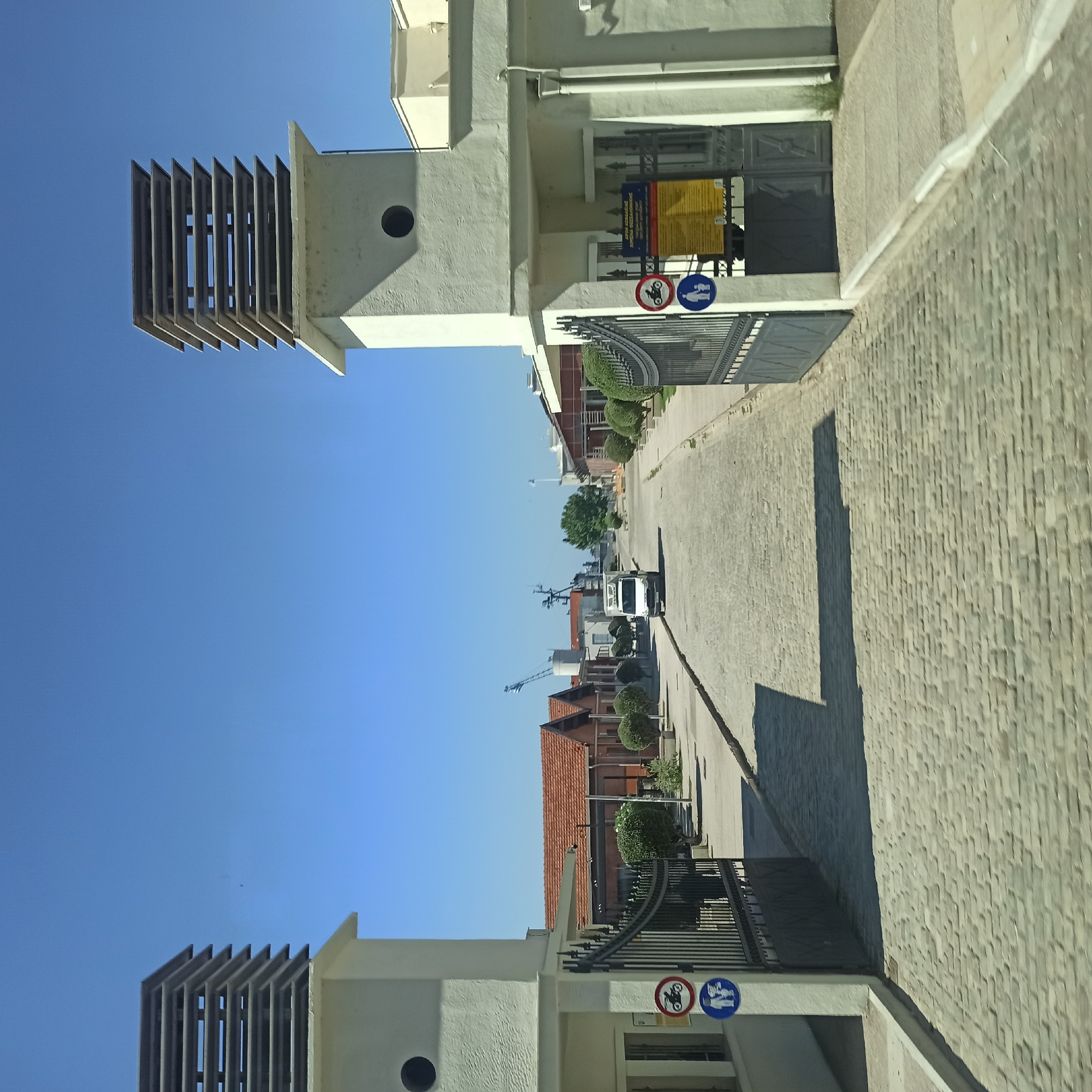The image depicts a sunlit day in an industrial or port area, likely within the city of Thessaloniki, Greece, given the presence of Greek text on a sign and the architectural style. A paved cobblestone road leads straight ahead into a complex lined with buildings that have terracotta-tiled roofs. On either side of the road, at the entrance to the complex, stand two identical white gatehouses topped with distinctive, stacked horizontal louvres. These gatehouses flank open wrought-iron gates.
Attached to the left gatehouse is a circular blue sign with a white motorcycle silhouette, indicating "no motorcycles." Below that is a blue circular sign with a white silhouette of two people, signifying "pedestrian crossing" or "pedestrian area." A similar set of signs is attached to the right gatehouse, though the specific details are less clear. A yellow sign with Greek text is also visible on the right gatehouse, likely containing information or regulations.
A white refrigerated truck is driving down the cobblestone road. In the background, construction cranes and parts of ships are visible, suggesting proximity to a harbor or shipyard. Green trees and bushes are strategically placed along the sides of the road, softening the industrial landscape. The sky is a clear, vibrant blue. The overall scene conveys a sense of order and activity within a functional, working environment.