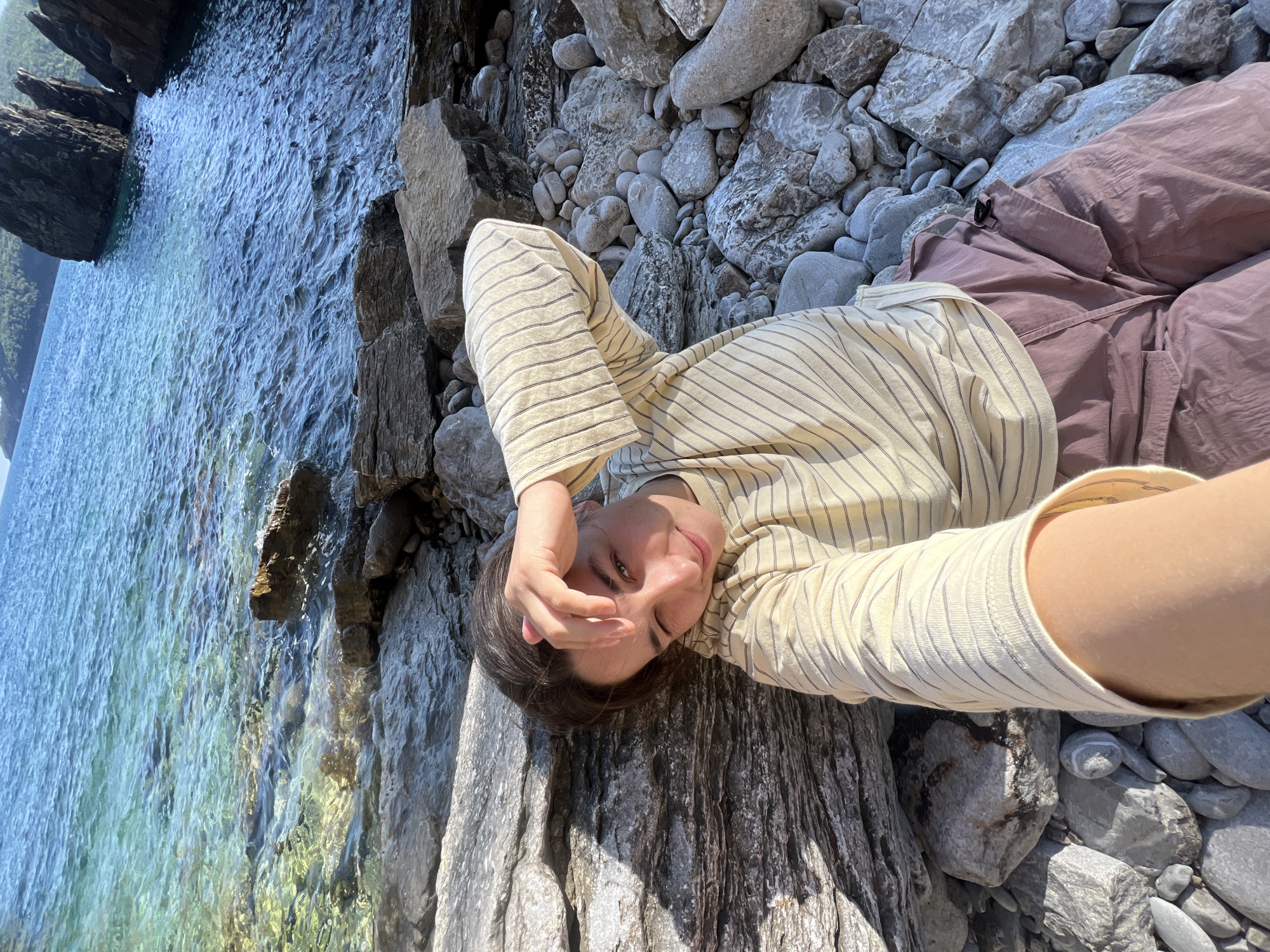 A young woman is lying on a large, grey rock alongside a body of water. She is wearing a light yellow, long-sleeved, striped shirt and mauve pants. She has dark hair and is holding her hand up to shield her face from the sun. The body of water is blue and clear, with rocks lining the edge. In the background, there are larger rocks and trees visible. The photo was likely taken in Boreti, Montenegro.