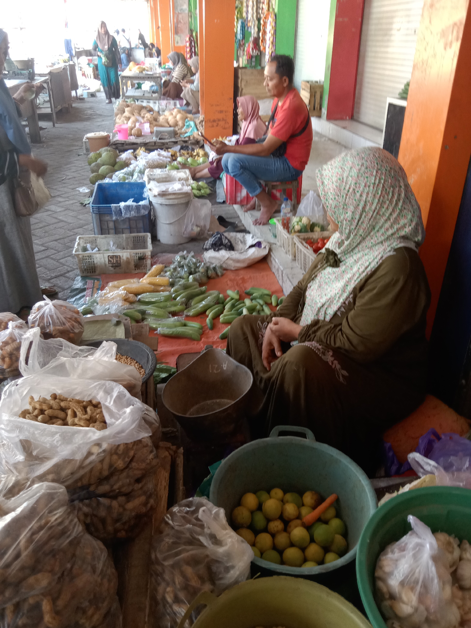 In a market stall in Sugihwaras, Indonesia, a woman wearing a headscarf sits on the ground, tending to produce. She is dressed in a long, olive-green garment with embroidered details. In front of her, on an orange tarpaulin, are piles of vegetables, including cucumbers, corn, and other items. Several plastic bags filled with nuts, possibly peanuts, are arranged on the ground. To her right, two large plastic buckets contain lemons and a carrot in one, and garlic in the other. In the background, other market-goers and stalls are visible, indicating a busy marketplace. The stalls are made of wood and corrugated metal, and the ground is paved with brick.