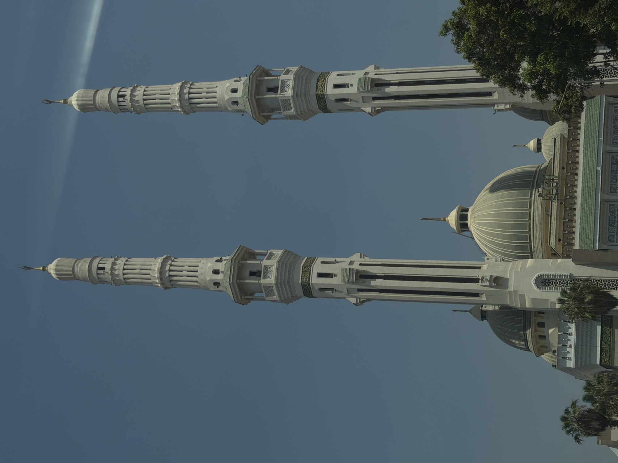 The image shows the Al-Azhar Mosque in Cairo, Egypt. The mosque features two prominent minarets that rise high into the clear blue sky. The minarets are tall and slender, with intricate architectural details and decorative elements. They have a light-colored stone facade, punctuated by rows of small windows and balconies. At the top of each minaret is a finial, topped with a crescent moon symbol. In the foreground, part of the mosque's domed structure is visible, as well as lush green trees. The sky is a vibrant blue with a thin, wispy cloud streaking across it. The overall scene depicts a grand religious structure against a bright, sunny backdrop.