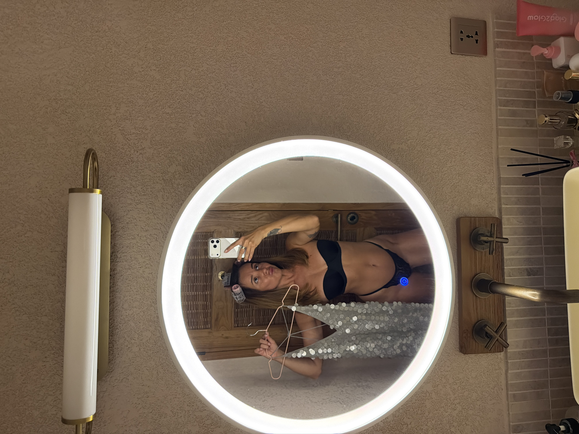 A woman is taking a mirror selfie in a bathroom, holding a sequined top on a hanger. She is wearing a black strapless bra and black thong, and has hair rollers in her hair. The bathroom features a large, circular, illuminated mirror, a modern faucet, and various toiletries on the counter. The wall has a textured finish. The scene appears to be in a hotel or resort bathroom in Mengwi, Indonesia.
