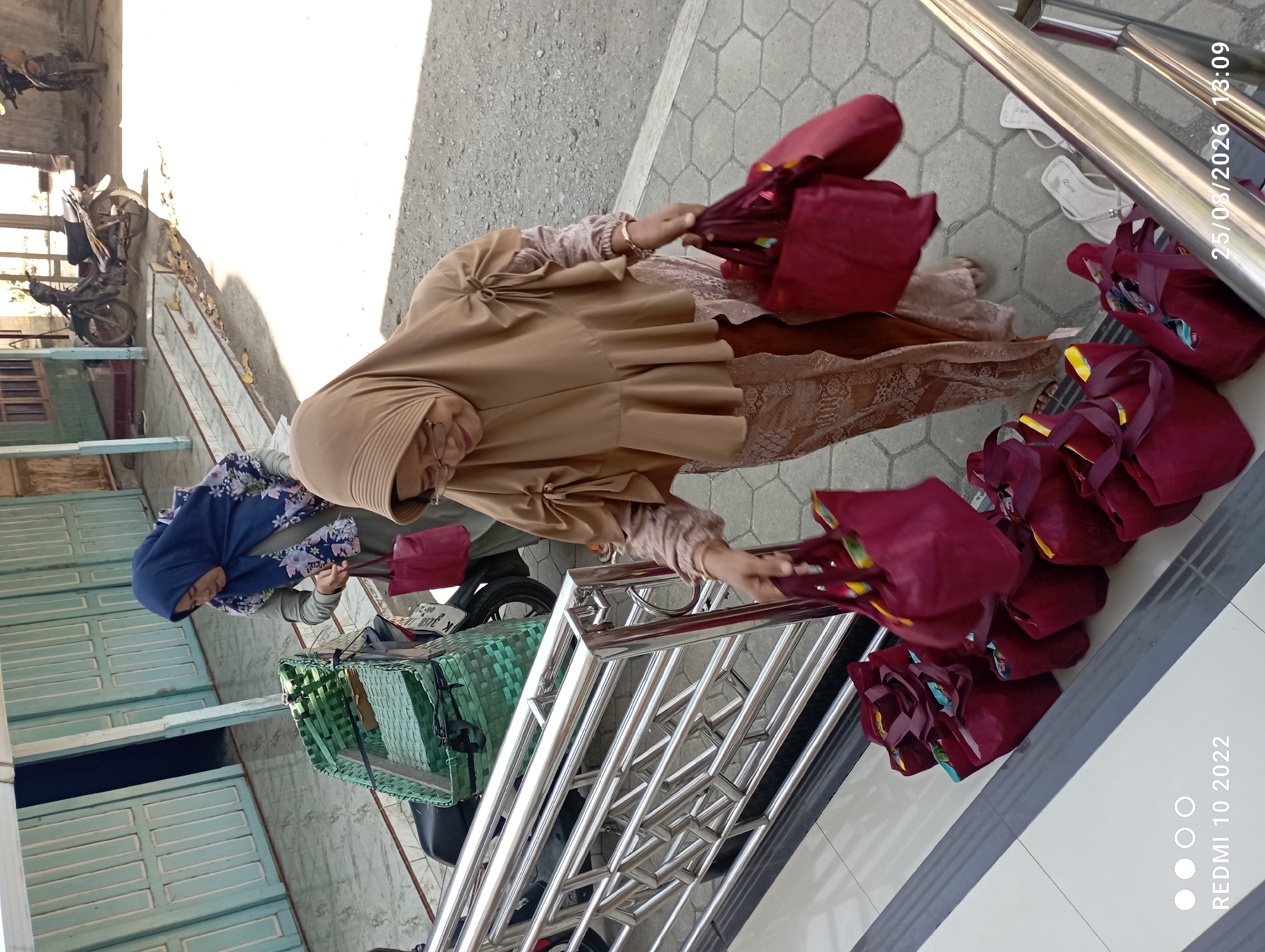 In Dukuhseti, Indonesia, two women in hijabs are standing near a motorcycle. One woman, wearing a blue hijab with floral patterns, is reaching into a green woven basket on the back of the motorcycle and holding a maroon bag. The second woman, wearing a tan hijab and glasses, is in the foreground, holding two maroon bags. In front of her, arranged on the tiled steps, are several more maroon bags. The environment appears to be outdoors, with paved ground and a building in the background. The time stamp on the image indicates it was taken on August 25, 2026, at 13:09.