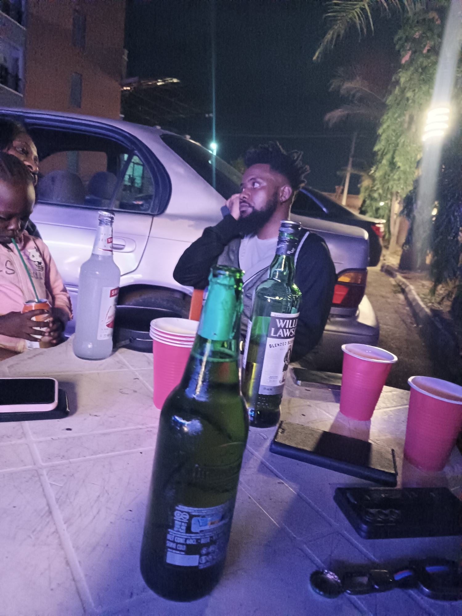 The image depicts a nighttime scene with a group of people gathered around a table. A man with a beard and dreadlocks is seated, seemingly on a phone call. A child, presumed to be female, sits to the left drinking from a beverage with a straw. Another person is partially visible behind the child. A silver sedan is parked nearby. The table is covered with a patterned white tablecloth and holds several items, including a Smirnoff Ice bottle, a green beer bottle, a bottle of William Lawson's Blended Scotch Whisky, several red plastic cups, and three smartphones. A bottle opener is on the table. The location appears to be outdoors, possibly a residential area, with a building visible in the background. The presence of cars and a streetlight implies an urban setting, possibly in Jos, Nigeria.