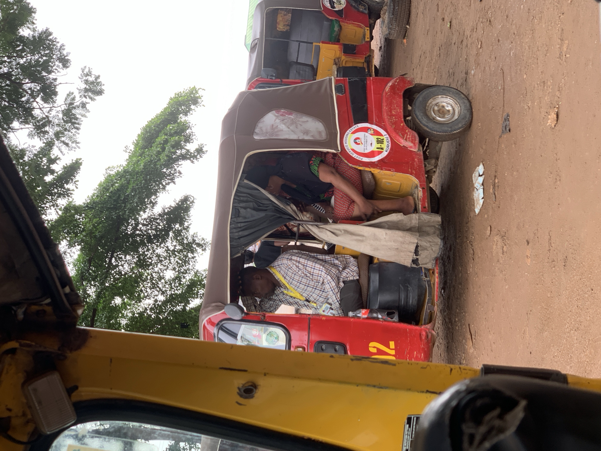 This media file captures a scene in Abuja, Nigeria, likely during the day as suggested by the bright overcast sky. The primary subjects are two auto-rickshaws, commonly known as "Keke napeps," parked side-by-side on a dirt road.
The auto-rickshaw in the foreground, which appears yellow, has its interior partially visible, including a rearview mirror and part of the dashboard. The auto-rickshaw in the background, predominantly red and yellow, is the focus of the activity. Inside this vehicle, three individuals are visible. One person, wearing a checkered shirt and a yellow lanyard, is slumped forward, seemingly asleep or resting. Another person, partially obscured, is dressed in a black top and patterned trousers, also appearing to be resting. A third individual is positioned further back in the auto-rickshaw. The vehicles have fabric canopies for shade.
Notable details include stickers on the red auto-rickshaw. One prominent circular sticker displays text and an image, including the words "POST SERVICE HOUSING ESTATE" and "A-102 KURUDA". Another yellow number "102" is visible on the side of the red auto-rickshaw. The ground is dusty and uneven, typical of unpaved roads. Lush green trees are visible in the background to the left, contrasting with the earthy tones of the road and vehicles. The overall impression is one of a busy transport hub or a roadside stop where drivers might be resting between journeys. The sky appears bright, indicating daylight, and the presence of overcast conditions might suggest it's not direct, harsh sunlight.