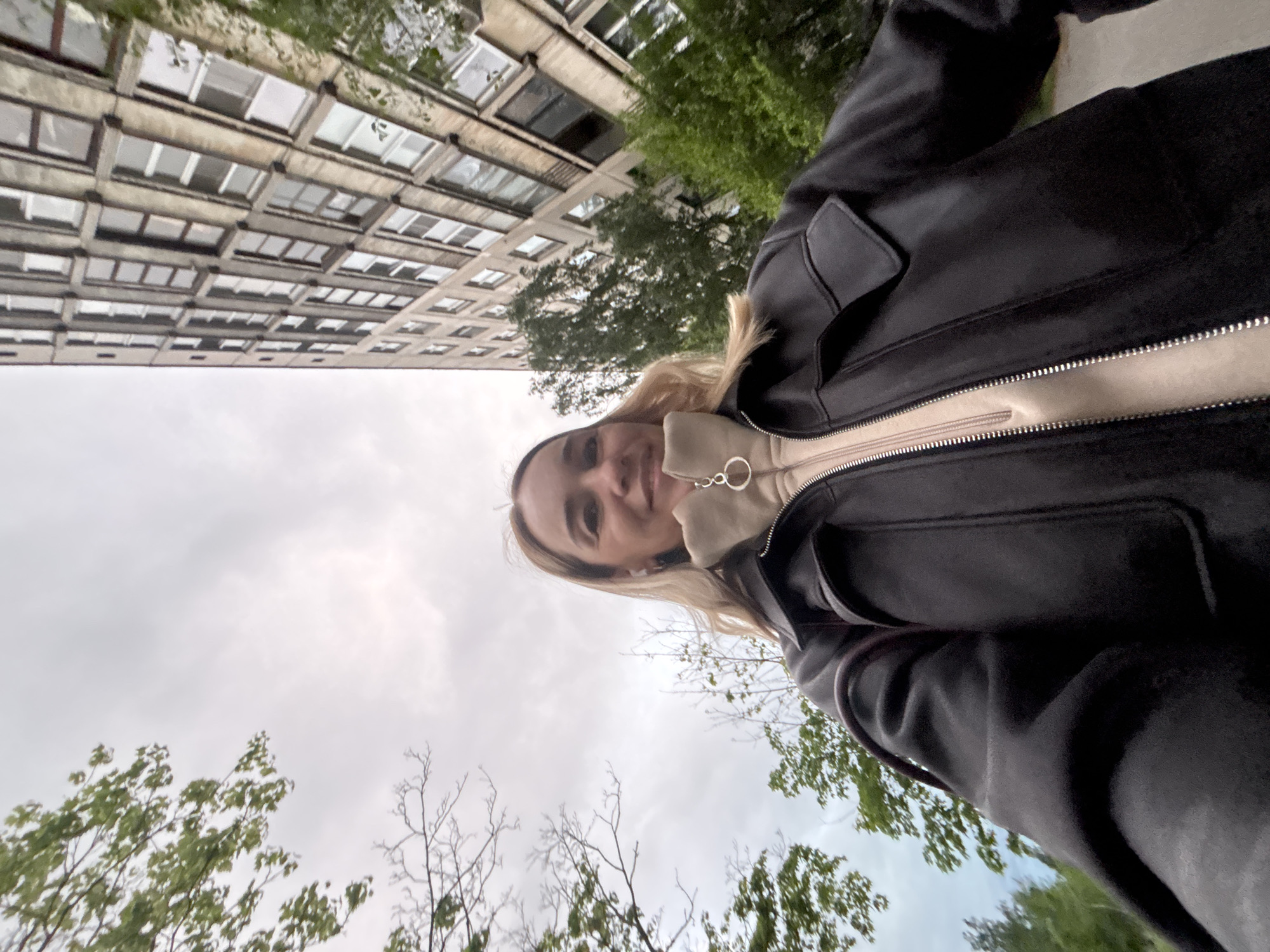 A young woman with blonde hair stands outside wearing a black jacket over a tan sweater. She is smiling slightly at the camera. Behind her is a large, grey, block-style building with many windows, partially obscured by green trees. The sky is overcast. The scene is in an urban area.