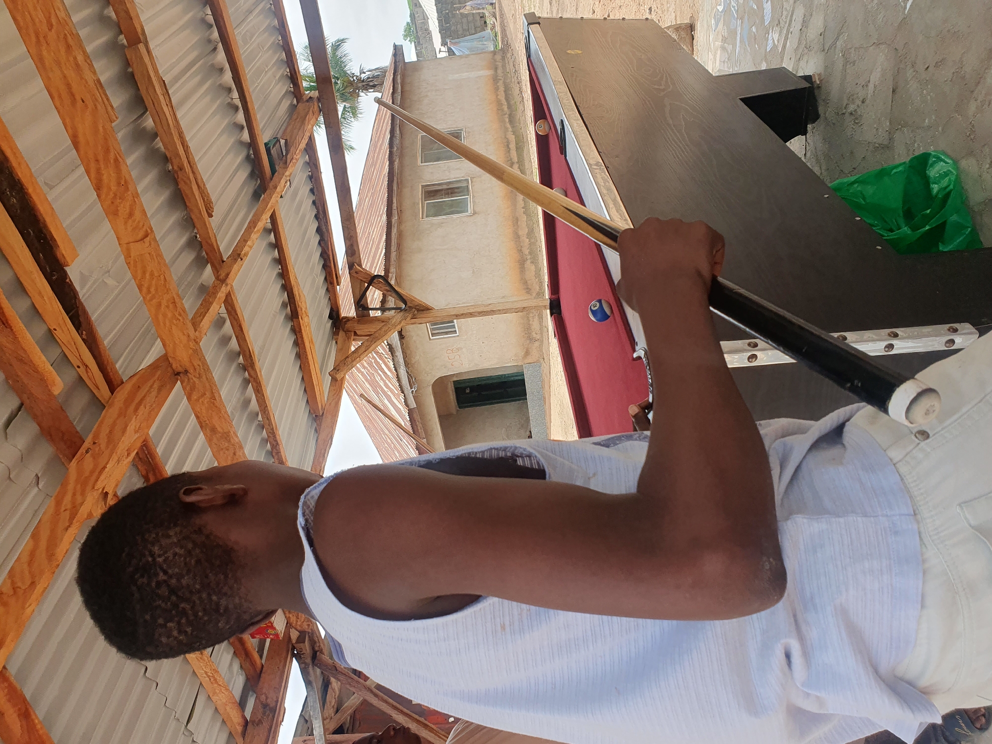 The image shows a young person playing billiards. The person is wearing a white tank top and white shorts. They are holding a pool cue and appear to be taking a shot. The pool table has a red felt surface and is partially covered by a dark wood table top. Several billiard balls are visible on the table.
The scene appears to be outdoors, under a makeshift shelter with a corrugated metal roof supported by wooden beams. In the background, there is a building with windows and a partially visible palm tree, suggesting a tropical or subtropical climate. The lighting suggests it is daytime, with the sun casting shadows.
Notable details include the unfinished nature of the shelter, indicated by the exposed wooden beams and metal roofing. The presence of a bright green plastic bag on the ground near the pool table might indicate casual storage or waste. There is no visible text in the image. The overall impression is one of a relaxed, informal setting for playing billiards, possibly in a rural or semi-urban area of Lokoja, Nigeria.