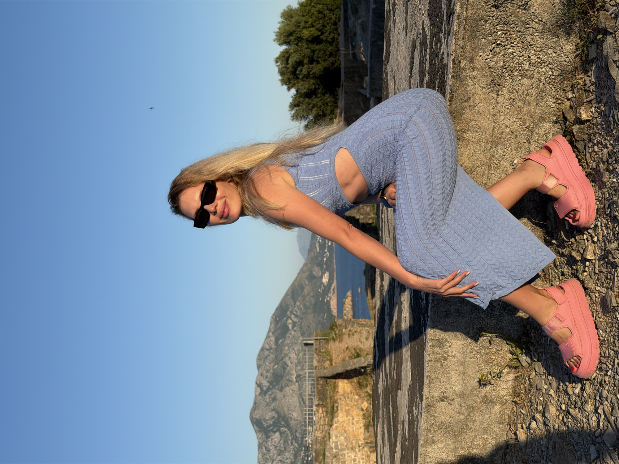 A blonde woman is seated on a weathered, light gray concrete surface outdoors during the golden hour, likely in Budva, Montenegro. She is turned slightly left, with her head angled towards the viewer, wearing black rectangular sunglasses and a light blue, sleeveless, knitted midi-dress with a cable pattern and side cut-outs. Her fingernails are painted light pink, and her toenails are red. She wears bright pink, chunky platform sandals with two straps across the foot and one around the ankle. A watch is visible on her left wrist. The ground directly around her consists of small stones and dirt. In the background, a large, rocky mountain range rises under a clear blue sky, with a body of water and a coastal town visible below the mountains. To the mid-left, old stone walls and structures with a metal railing are present. To the right background, a dense cluster of dark green trees grows on higher ground. A small, dark bird is visible in the upper right sky. Long shadows are cast from the left.
