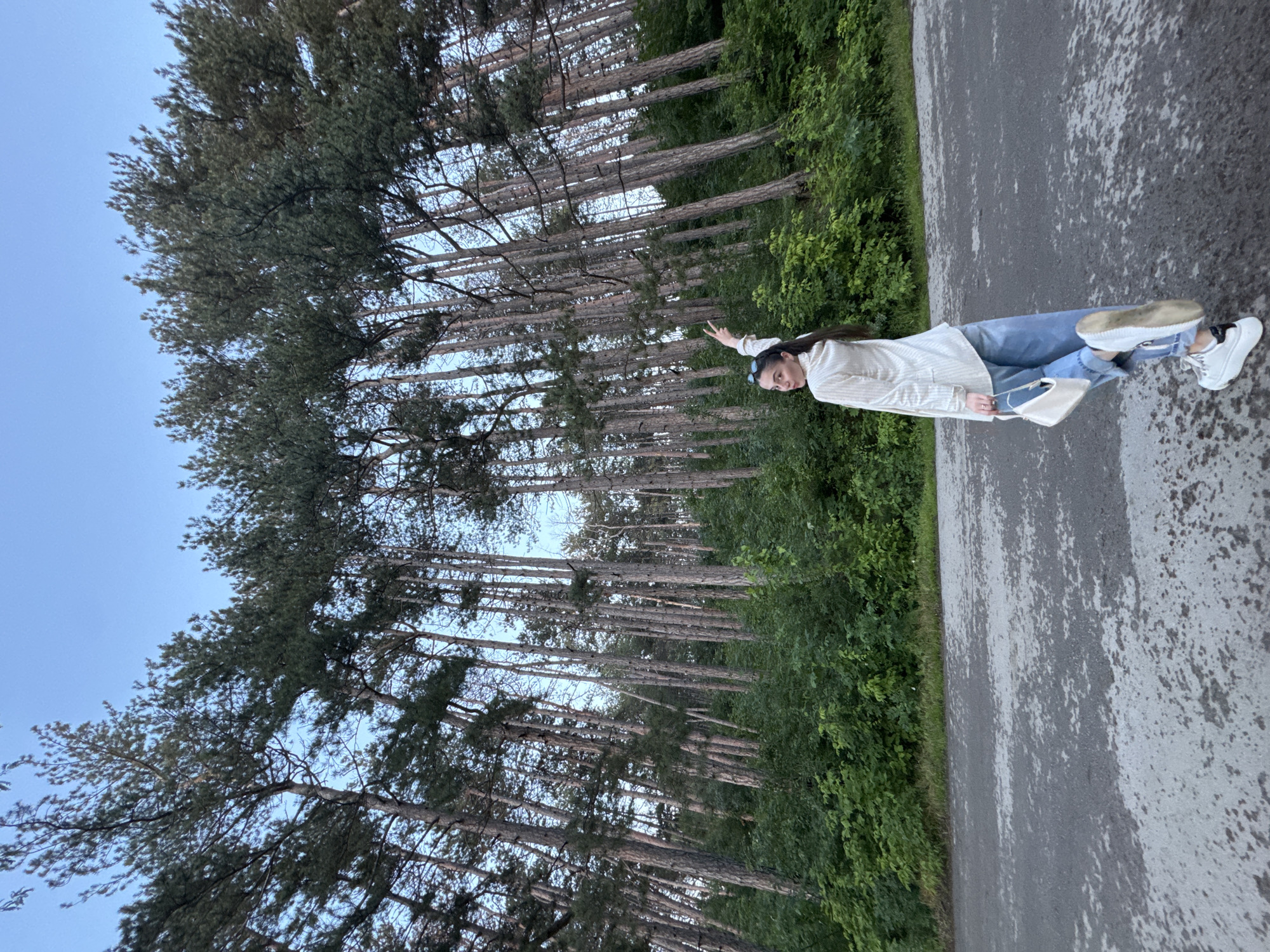 This low-angle photograph depicts a young woman posing on an asphalt road adjacent to a dense forest in Targovishte, Bulgaria. The woman, with dark hair pulled back and sunglasses on her head, wears a light-colored, vertically striped long-sleeved shirt, loose-fitting light blue jeans, and white platform sneakers with black heel accents. She is positioned facing away from the camera but looks back over her left shoulder with a slight smile. Her right arm is raised, forming a 'peace' sign with her index and middle fingers, while her left hand holds a small white rectangular shoulder bag. Her left leg is partially lifted, exposing the sole of her sneaker. The background features a multitude of tall, slender evergreen trees, likely pines, with light-colored trunks and dense dark green canopies. A thick layer of green undergrowth and bushes covers the forest floor. A clear, light blue sky is visible above the treetops. The grey asphalt road surface in the foreground and mid-ground shows irregular lighter grey patches.