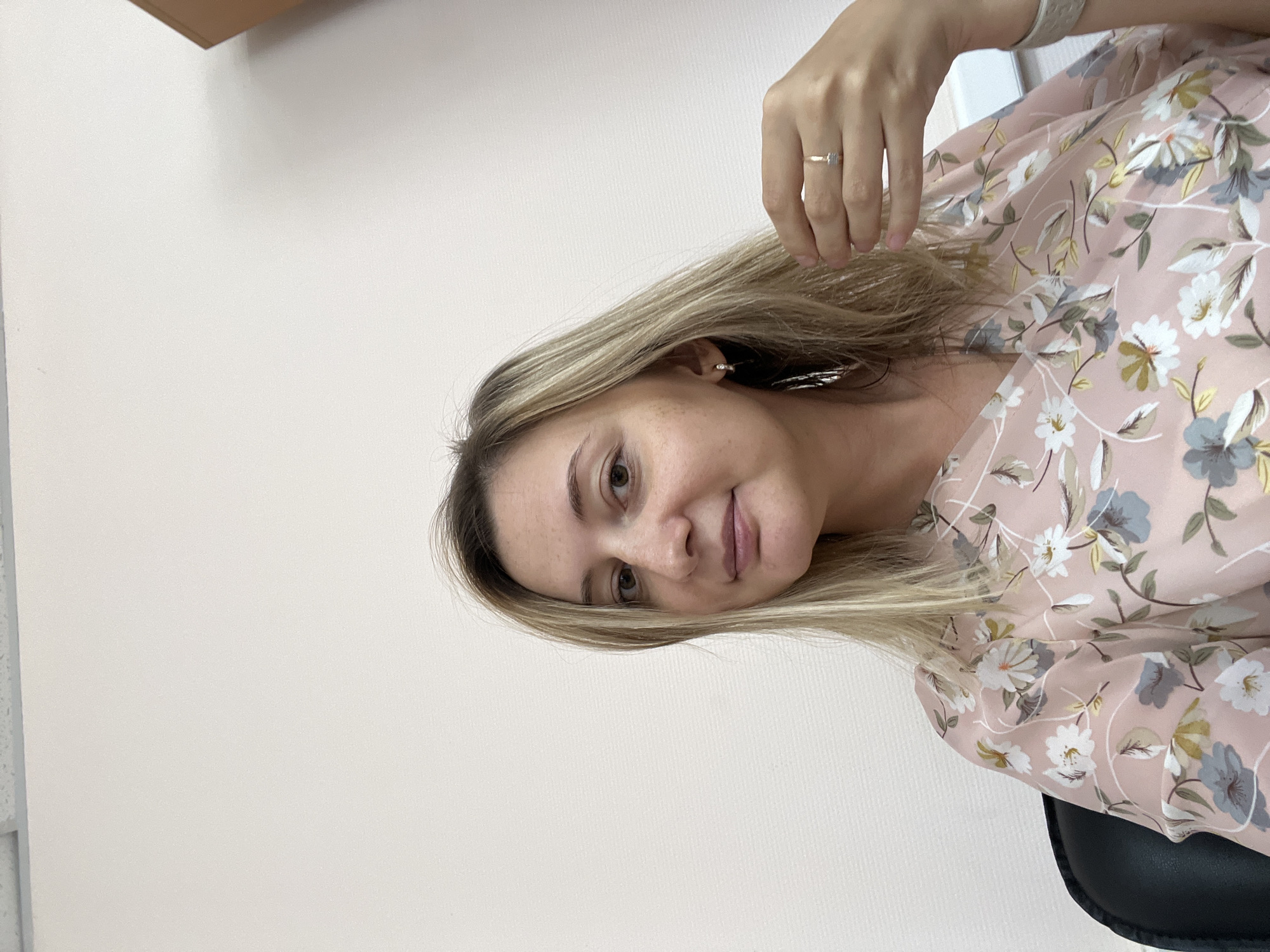 A medium shot captures a woman with blonde hair and fair skin from the chest up, seated in a black chair. She is looking towards the camera, her head slightly tilted. The woman is wearing a pink blouse with a floral pattern featuring white, blue, and yellow flowers. She has on small hoop earrings and a ring on her right ring finger. Her right hand is raised, touching her hair near her ear. The background is a plain, light-colored wall. No specific location in Volgograd, Russia, can be identified from the visual information provided.