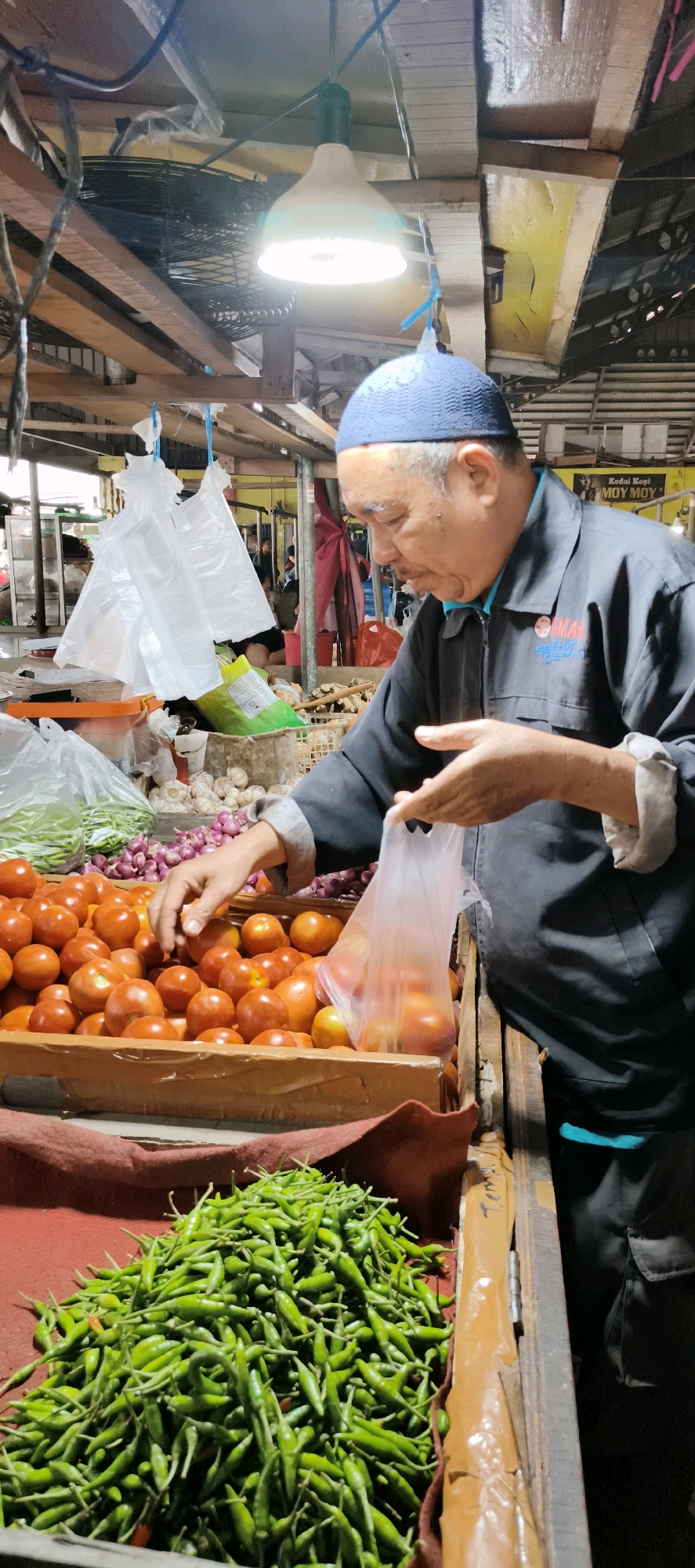 A man wearing a blue kufi and a dark jacket is seen at a market stall in Jambi City, Indonesia. He is holding a transparent plastic bag filled with tomatoes, appearing to be in the process of selecting or bagging them. In front of him, a wooden crate overflows with bright red tomatoes, and to the left, there are more tomatoes in a wooden tray. To the right of the tomatoes, a pile of green chili peppers is visible on a red cloth. In the background, other market goods and shoppers can be discerned. The stall is covered with a wooden structure, and a bright light hangs from the ceiling, illuminating the scene.