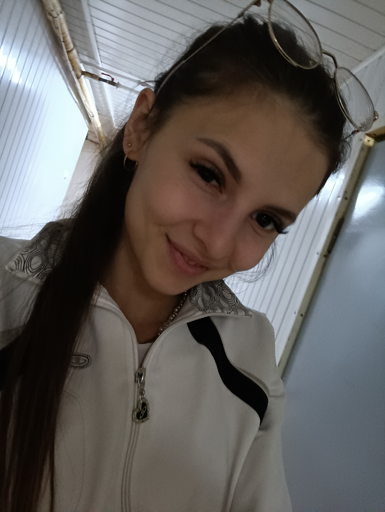 A young woman is pictured indoors. Her dark hair is partially pulled back, and glasses sit on her head. She wears a white jacket with black accents and a decorative zipper pull. The background shows a white paneled wall and ceiling, suggesting a utilitarian or commercial space. While I am unable to pinpoint an exact location in Прикамский, Russia, I can tell you that she is indoors.