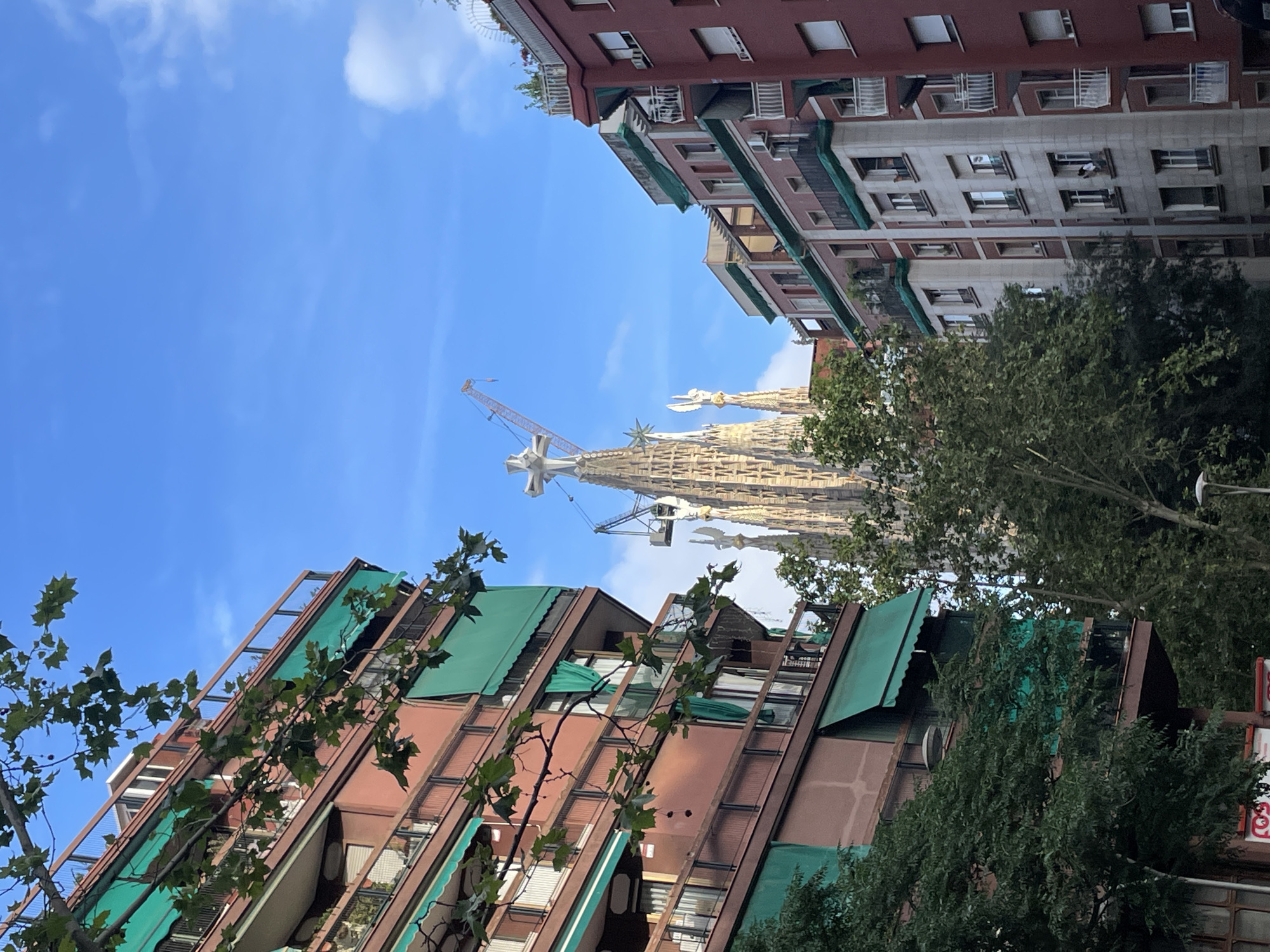 This is a street-level, eye-level shot of the Sagrada Familia Basilica in Barcelona, Spain. The Basilica's iconic spires are visible in the background, peeking above the surrounding apartment buildings. In the foreground, a large crane, likely used for ongoing construction on the Basilica, is visible against the blue sky. To the left of the frame, a brown apartment building with green awnings and balconies dominates the scene. To the right, another apartment building with a red facade and white balconies is partially visible. Lush green trees line the street, adding a touch of nature to the urban landscape. The sky is a vibrant blue with a few wispy clouds.