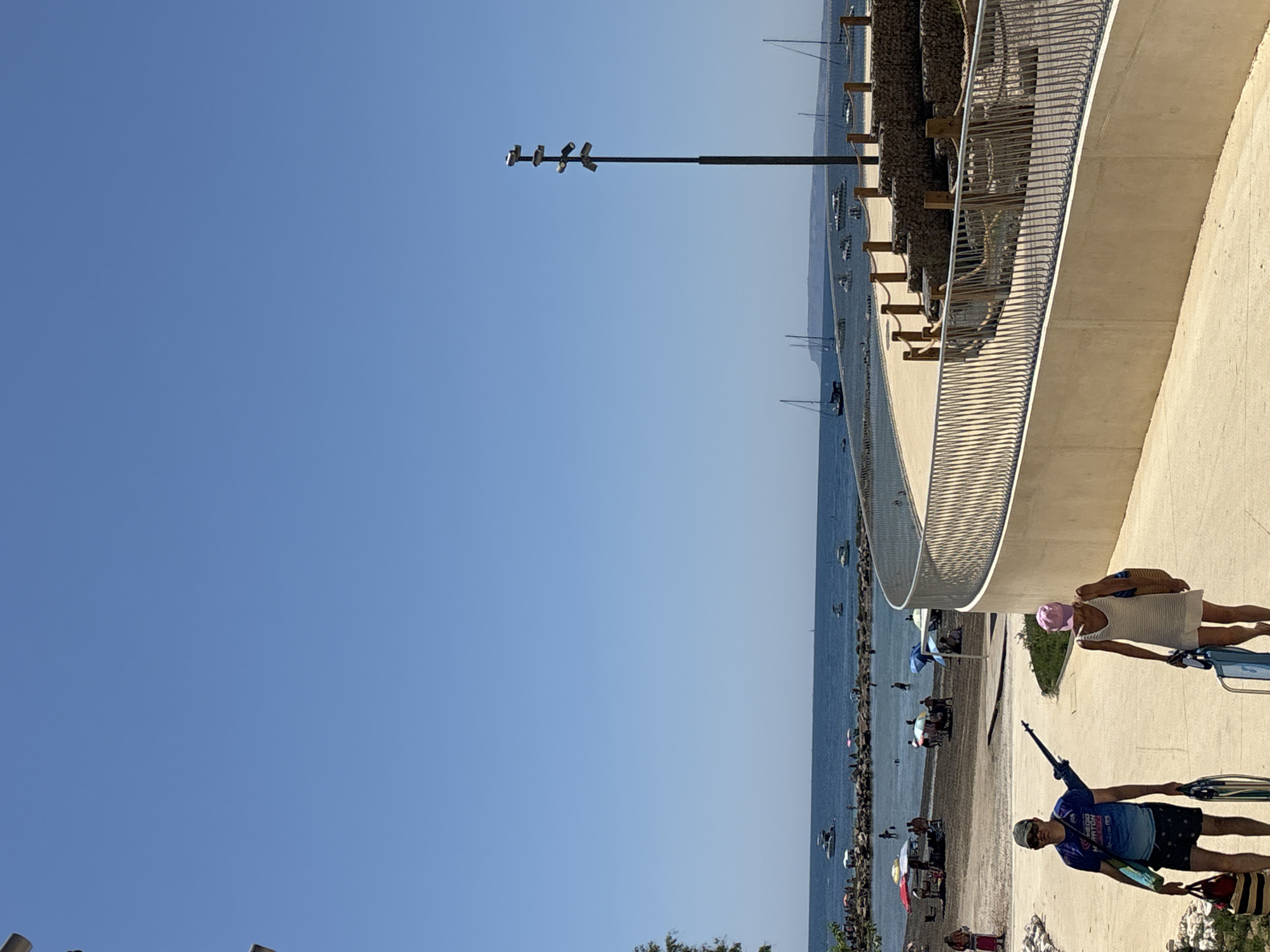 This image depicts a sunny day at a beach area, likely in Alicante, Spain, with a clear blue sky dominating the upper half of the frame. In the foreground, a man wearing sunglasses, a blue and purple t-shirt with text, and patterned shorts walks away from the camera, carrying a tote bag and a folded chair. Beside him, a woman in a white dress and pink hat also walks away from the camera, carrying a straw tote bag and a blue folded chair. They are on a paved walkway that curves around a landscaped area with stone terracing and metal railings.
In the mid-ground, a bustling beach scene unfolds. Numerous people are spread across the sand, some sitting under colorful umbrellas, while others are in the water. Several small boats are anchored in the shallow, calm sea. Further out, more boats, including sailboats, are visible on the horizon. A tall lamppost with multiple lights stands prominently on the right side of the image, near the elevated walkway. The overall atmosphere is one of a relaxed summer day at a coastal resort.