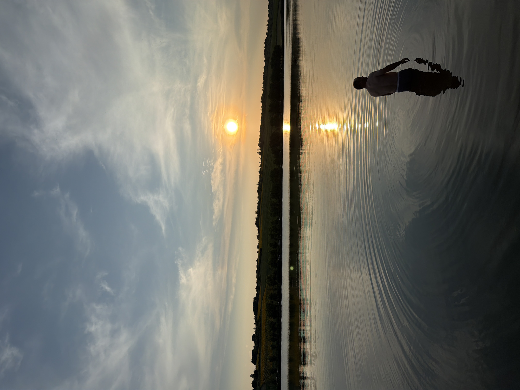 At sunset, a shirtless man stands waist-deep in a calm lake, facing away from the camera. The sun, a bright orange orb, is positioned in the upper right quadrant of the sky, casting a shimmering reflection on the water's surface. The lake is surrounded by a tree-lined shore, with rolling hills and fields in the background, suggesting a rural setting. The sky is a mix of blue and wispy white clouds, with gradients of orange and yellow near the horizon, indicative of the setting sun. The man's body is silhouetted against the water, and his reflection is visible below him, disturbed by subtle ripples. The image captures a serene moment in nature, likely during twilight hours.