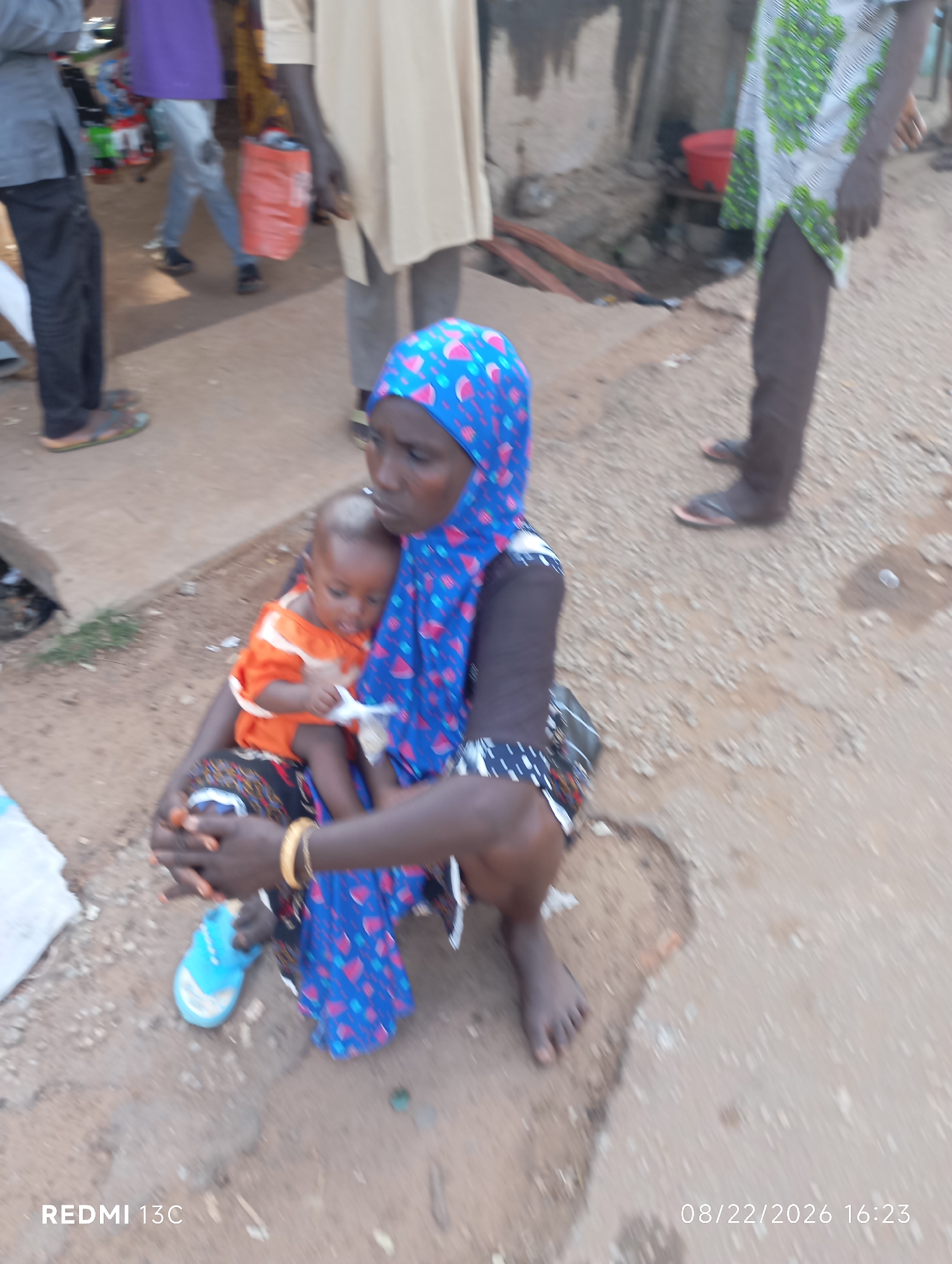 A slightly blurred, eye-level medium shot shows a woman in a blue hijab with pink and white patterns holding a baby. The woman's arms are wrapped around the baby, who is wearing an orange shirt and appears to be reaching for something held by the woman. The woman has dark skin, and is wearing a gold bracelet on her right wrist. Her legs are visible, with a blue sandal on her left foot and her bare right foot on the ground. The background is out of focus, but shows a dirt ground with some scattered debris, and the legs of other individuals in the background. A red plastic bag is visible in the background, along with parts of clothing worn by other people. The timestamp "08/22/2026 16:23" and "REDMI 13C" are overlaid on the bottom right and bottom left of the image, respectively. The scene appears to be outdoors, possibly in a marketplace or public area, in Shendam, Nigeria.