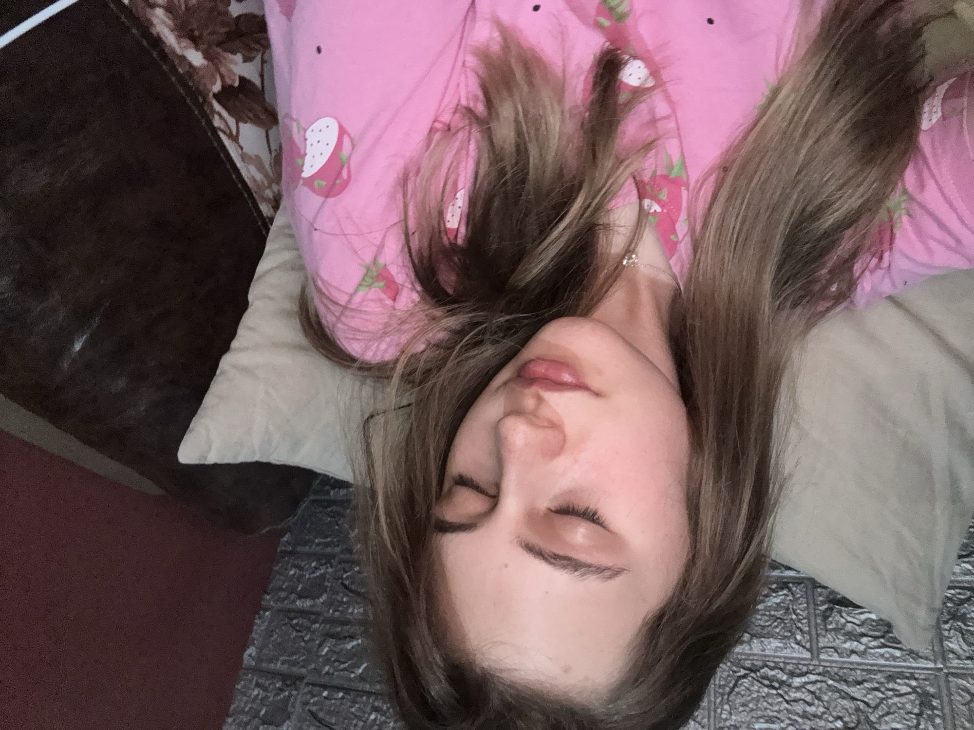 A young woman is lying on a pillow in a supine position. She is wearing pink pajamas with a strawberry pattern. Her eyes are closed. She has light brown hair. The background includes a brown object to the left and a patterned gray surface to the right. The location is unspecified and unidentifiable as Panagyurishte, Bulgaria, given the available visual information.