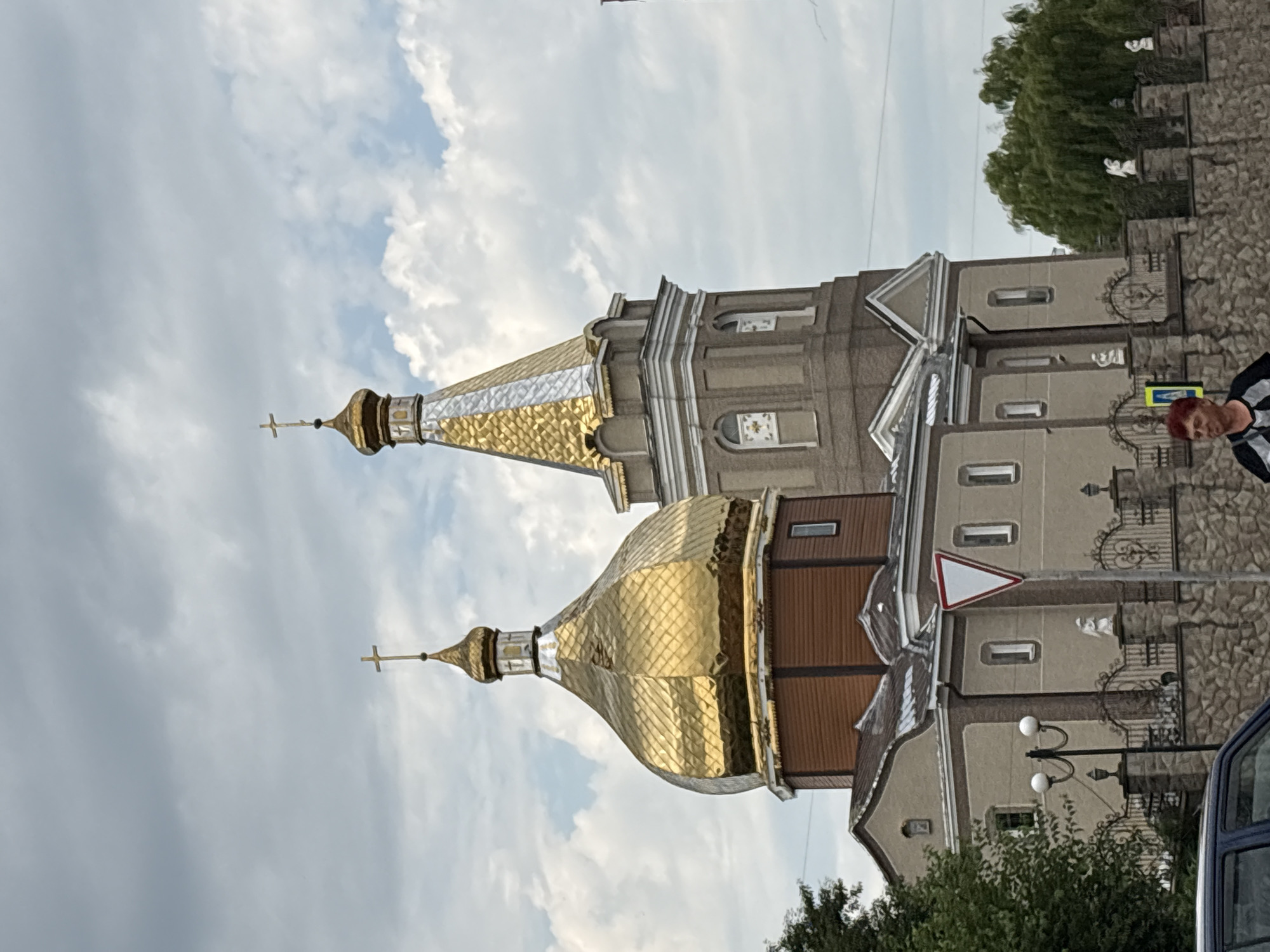 The image shows a church with golden domes and a bell tower, situated in the city of Lanivtsi, Ukraine. The architecture is traditional for Eastern Orthodox churches, featuring multiple domes, with the prominent ones being golden and silver. The church is made of brick and stone, with a light-colored facade. In front of the church, there is a stone wall and an ornate metal fence. A street sign is visible near the fence, and a traffic light can be seen to the left of the church. A woman with short red hair is walking in the foreground, facing the camera. The sky is overcast with clouds. The image appears to have been taken during the daytime, likely in the summer or spring given the foliage.