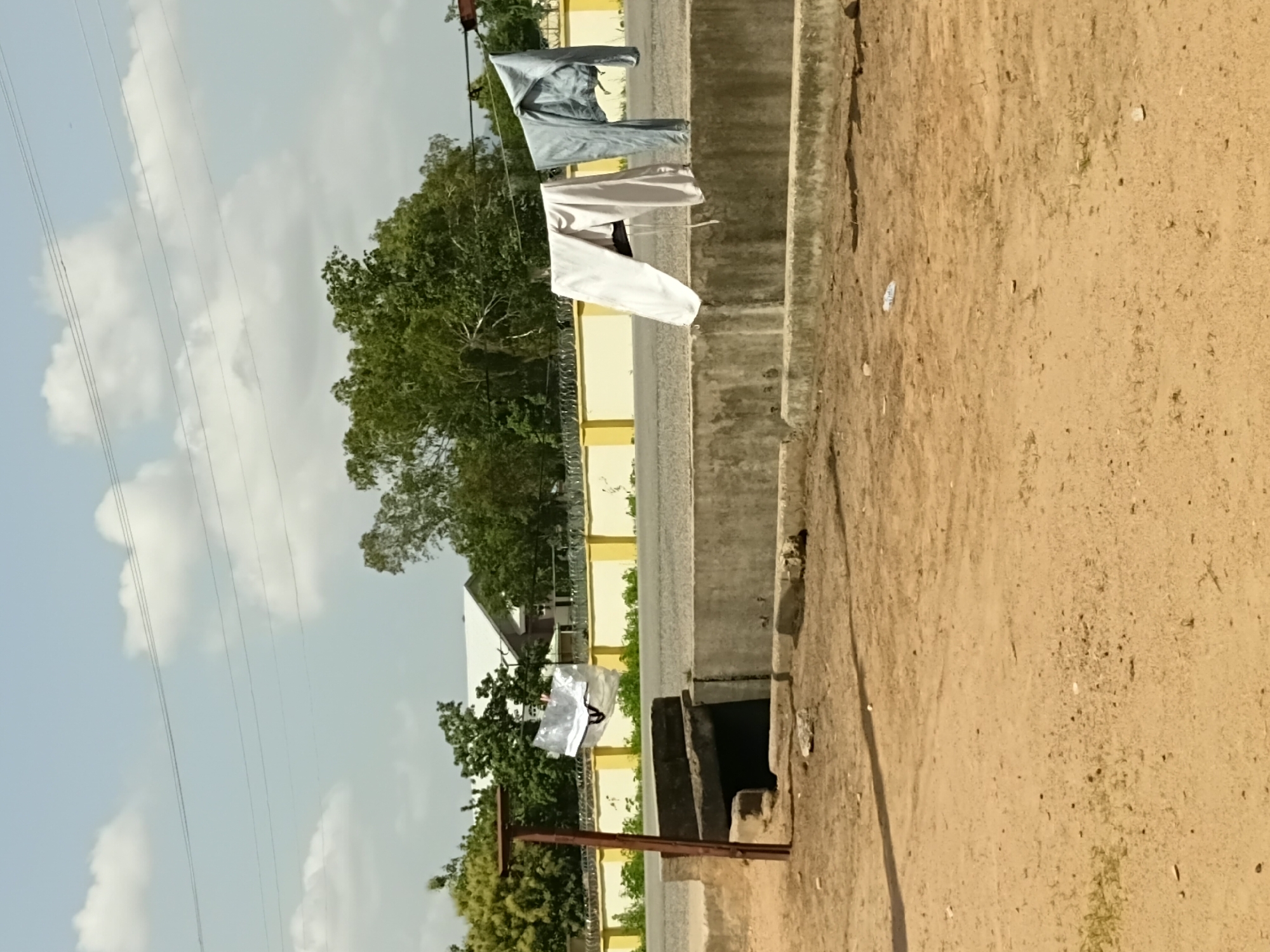 In Jalingo, Nigeria, laundry hangs from a clothesline strung across a rural road. The scene is set during the day, under a bright, partly cloudy sky. Clothes, including a grey shirt and white pants, are draped on the line, gently swaying in the breeze. The road runs parallel to a dirt embankment, which is the foreground of the image. Beyond the road, a line of yellow buildings is visible, interspersed with lush green trees. Power lines crisscross the sky, indicating the presence of infrastructure. The overall impression is one of everyday life in a Nigerian town, with laundry being air-dried under the African sun. The image is oriented vertically.