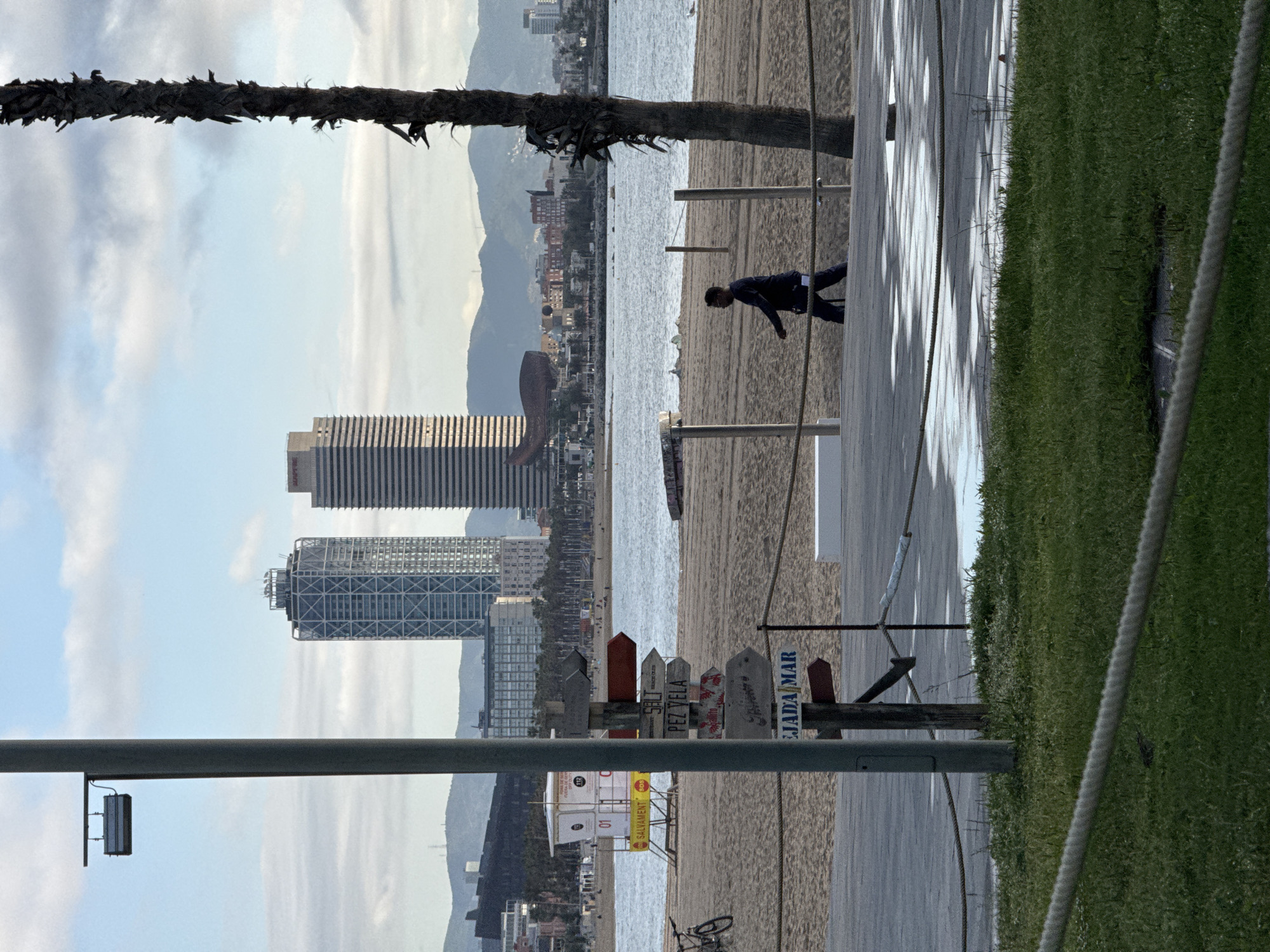 A person walks along a wooden boardwalk at Barceloneta Beach in Barcelona, Spain. The twin towers, Hotel Arts and Torre Mapfre, stand prominently in the cityscape. The beach is visible, with the sea and mountains in the background. A palm tree trunk is in the foreground, and a sign with the name "CIUTADA MAR" can be seen. There is grass to the right, with a rope defining its edge. There is a bicycle on the sand and a lifeguard station visible.