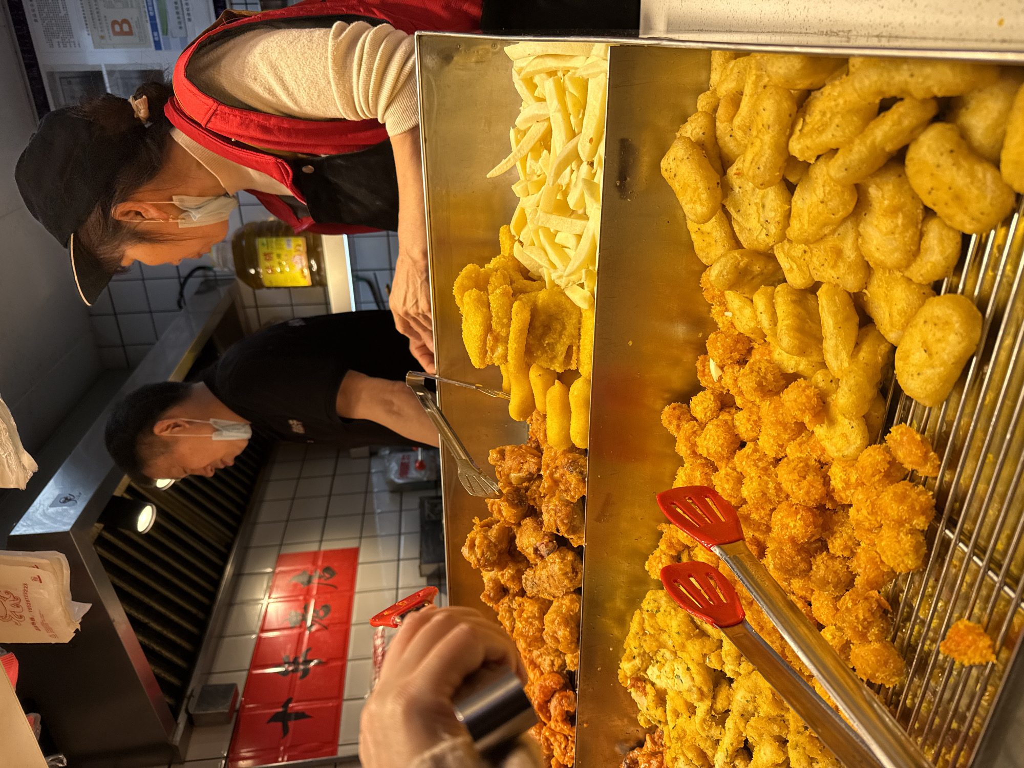 In a commercial kitchen, likely in Liangjiang, China, two workers wearing face masks are tending to a food preparation area. A woman with a black cap and red vest is near a container divided into sections filled with various fried foods: french fries, fried dough sticks, and golden-brown nuggets. Another worker, a man in a black shirt and face mask, stands in the background. In the foreground, a hand holding metal tongs reaches toward the food. A red banner with black Chinese characters hangs on the tiled wall beside the ventilation hood.