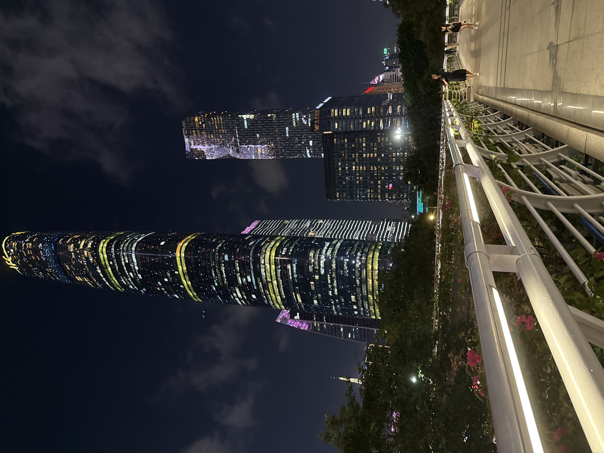 This is a nighttime scene of the Tianhe District, China skyline from a pedestrian overpass. Prominent in the image are illuminated skyscrapers. One building is cylindrical, featuring horizontal bands of light across its facade. Another, taller building has a rectangular form.
Two people are on the overpass. A woman in a dark dress stands near the railing, while another person walks further along the overpass. A white metal railing with floral decorations lines the side of the overpass, separating the sidewalk from the view of the city. The sky is dark with scattered clouds.