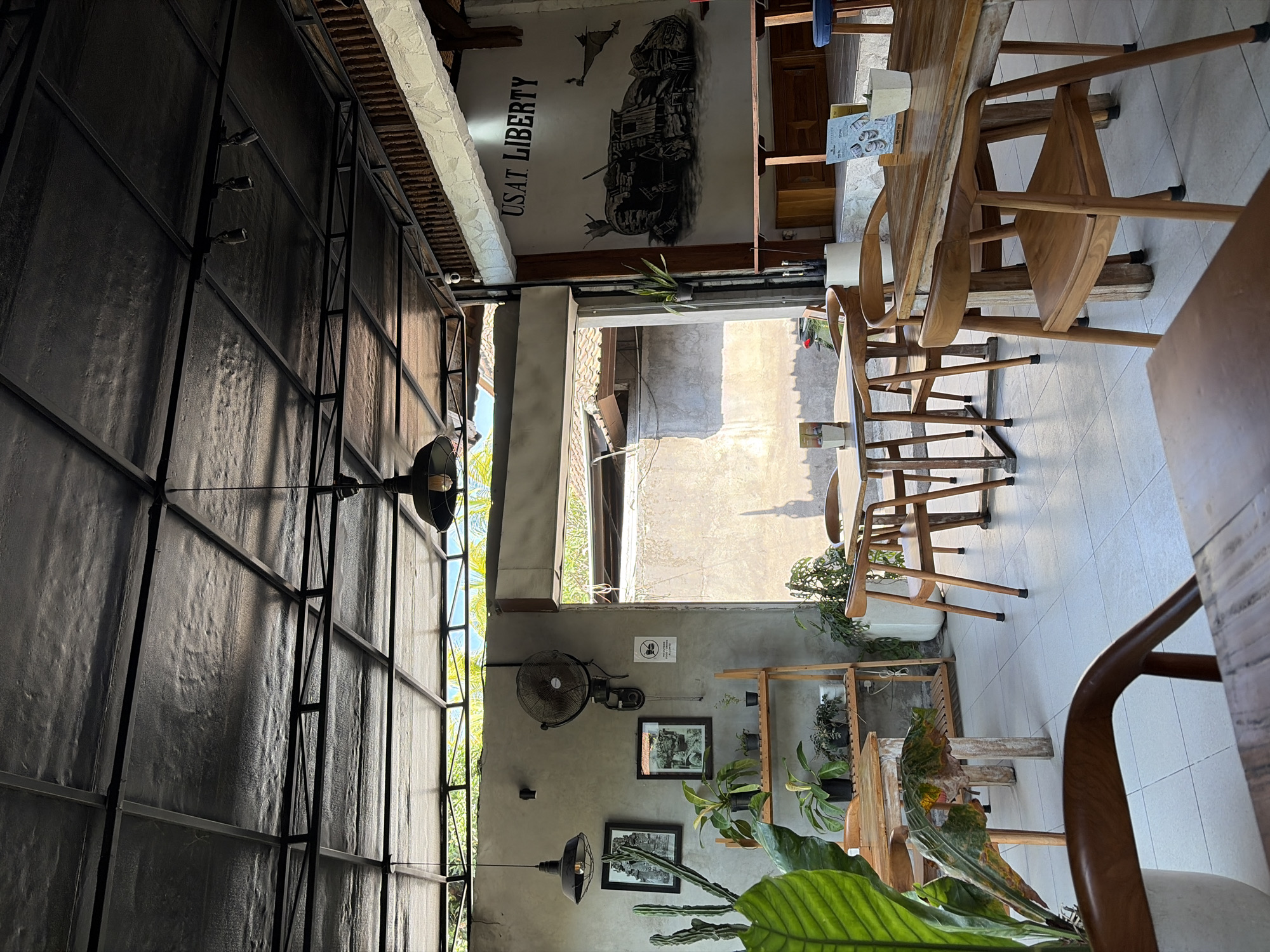 The image depicts the interior of a cafe, likely in Kubu, Indonesia, with an open-air section. The cafe features a rustic aesthetic with a dark metal ceiling structure and exposed beams. Wooden tables and chairs are arranged on a tiled floor.
On the left wall, there are two framed pictures hanging, a large oscillating fan, and a shelf unit filled with potted plants. To the right, a mural is painted on the wall, depicting a stylized vehicle and the text "USAT. LIBERTY". A wooden counter or bar is visible in front of the mural.
Natural light streams in from the open doorway and windows, creating shadows on the floor and walls. The environment appears to be a casual dining or seating area, possibly an outdoor cafe or a restaurant with an open-air design. There are no people visible in the image.