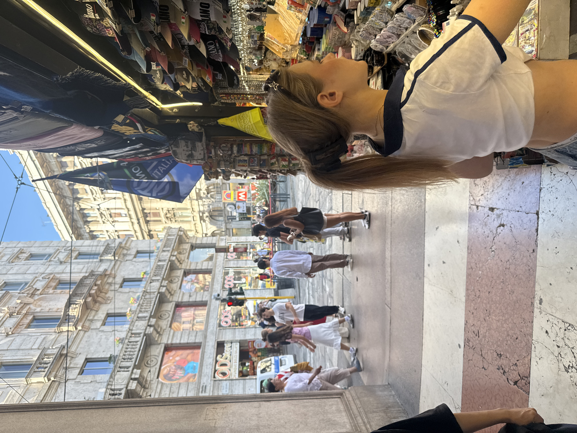 The image captures a street scene in Milan, Italy, likely near the Duomo, given the architectural style of the buildings and the presence of Italian flags and merchandise. In the foreground, a young woman with blonde hair pulled back in a ponytail is seen from the side, wearing a white cropped shirt with navy trim and denim shorts. She is facing a stall displaying various souvenirs, including hats and flags.
Behind her, a street unfolds with pedestrians walking on the sidewalk. Several individuals are visible, some dressed casually in t-shirts and shorts, while others are in more formal attire. One person is wearing a white long-sleeved shirt and brown pants, and another is in a white short-sleeved shirt and black pants. A traffic light with a red signal is visible further down the street. The buildings on either side of the street exhibit ornate Italian architecture, with balconies and detailed facades. The overall atmosphere suggests a bustling urban environment on a sunny day.
