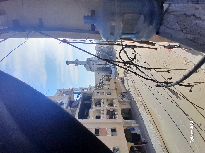 Damaged buildings line a street in a war-torn area. A mosque minaret is visible in the distance. Debris litters the scene. A water bottle sits in the upper right of the frame. Power lines crisscross the sky. The image is taken from an elevated perspective.