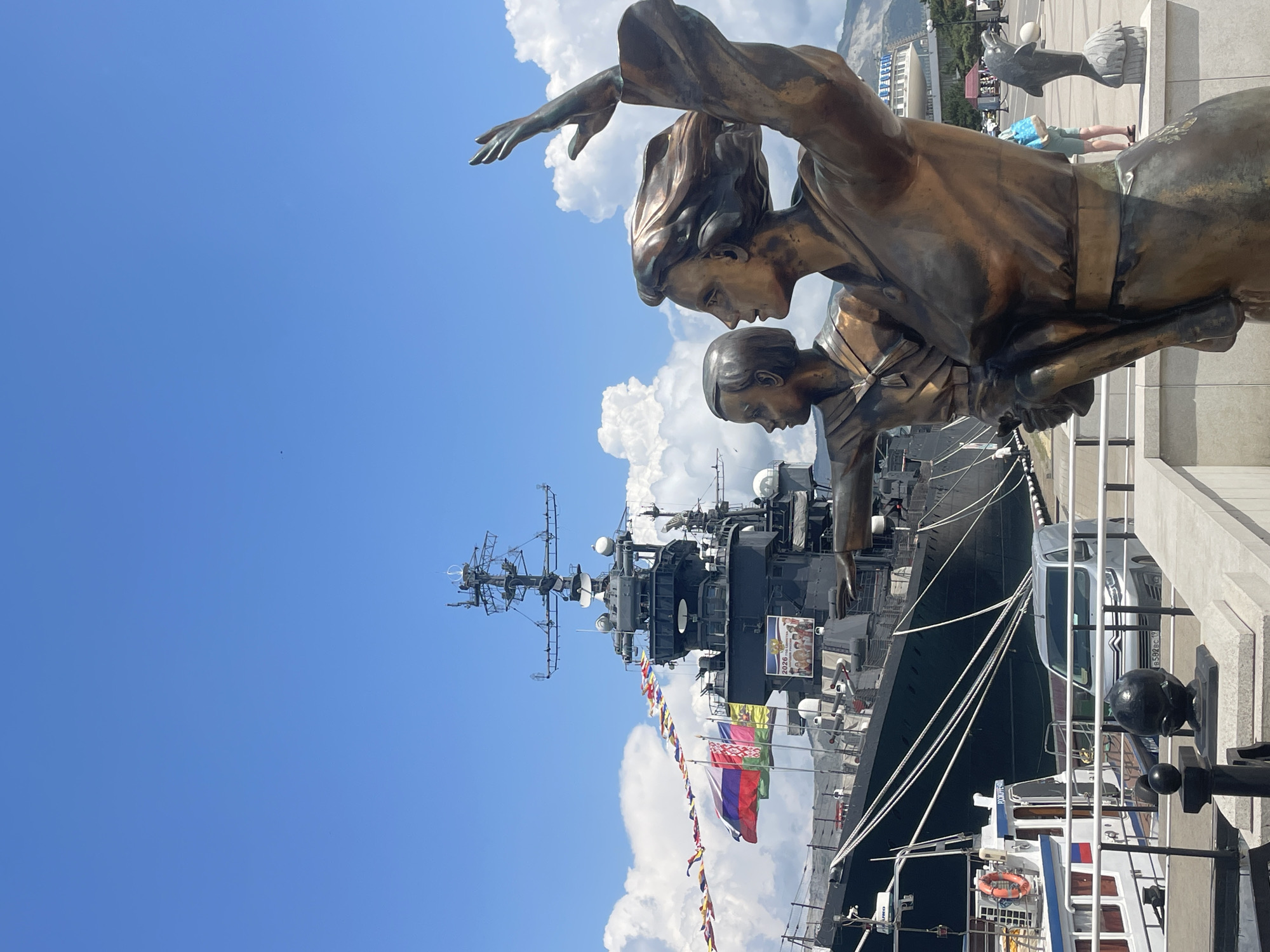 A bronze statue depicting a woman carrying a child is in the foreground of the image, positioned on a concrete embankment overlooking a harbor in Novorossiysk, Russia. The woman is looking to the right, with her right arm raised and hand open, while the child, held in her left arm, points forward towards a large, dark gray warship docked behind them. The warship has numerous antennas and turrets visible, and flags, including the Russian flag, are flying from its mast.
Several other boats are moored alongside the warship, and a silver van is parked on the quay. In the background, there is a white building with a rounded roof, and a mountain range is visible beyond that. The sky is blue with scattered white clouds, and the overall scene suggests a sunny day at the harbor. The statue and warship are likely part of a memorial or museum.