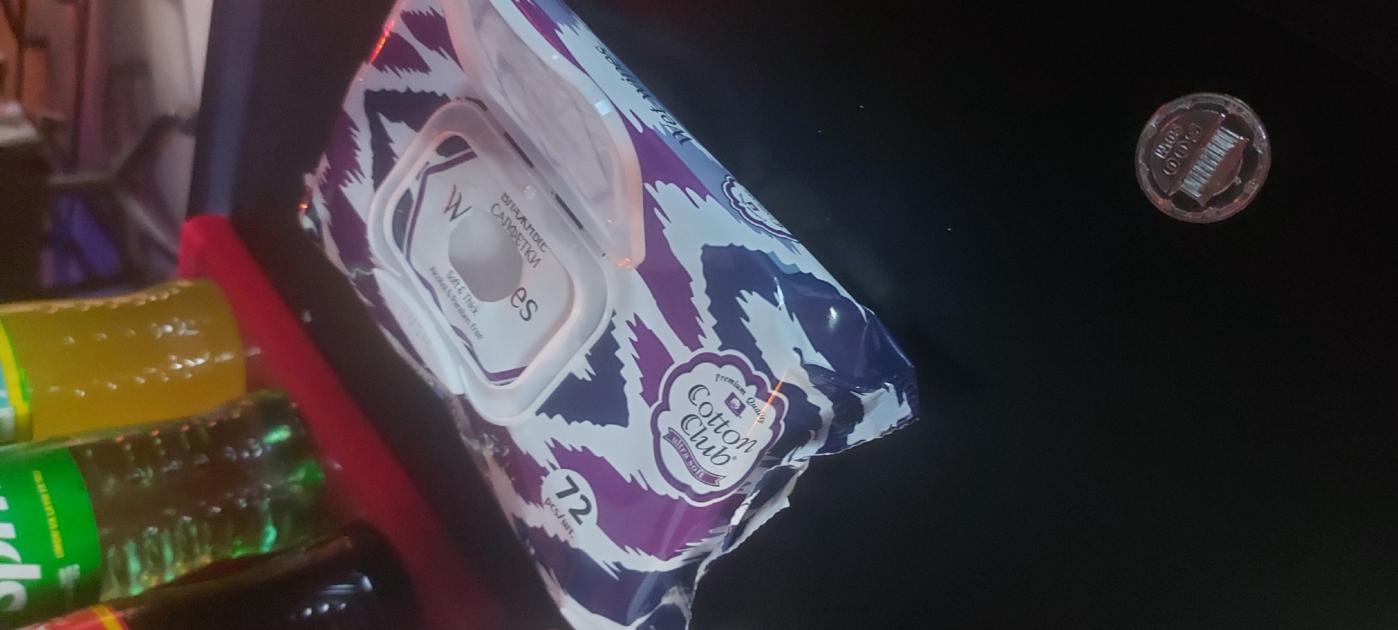 The image shows a pack of Cotton Club wet wipes, featuring a purple and white patterned design, positioned near a bottle of Sprite. The wet wipes' label includes text in both English and Russian, indicating "Soft & Thick" and "Alcohol & Paraben free." The pack contains 72 wipes. A round sticker with a barcode is visible in the upper right corner of the frame. Based on the Russian text on the wipes, the location is likely in a region where Russian is spoken, and given the presence of the product brand and other details, it is likely in a market or store within the city of Tashkent, Uzbekistan.