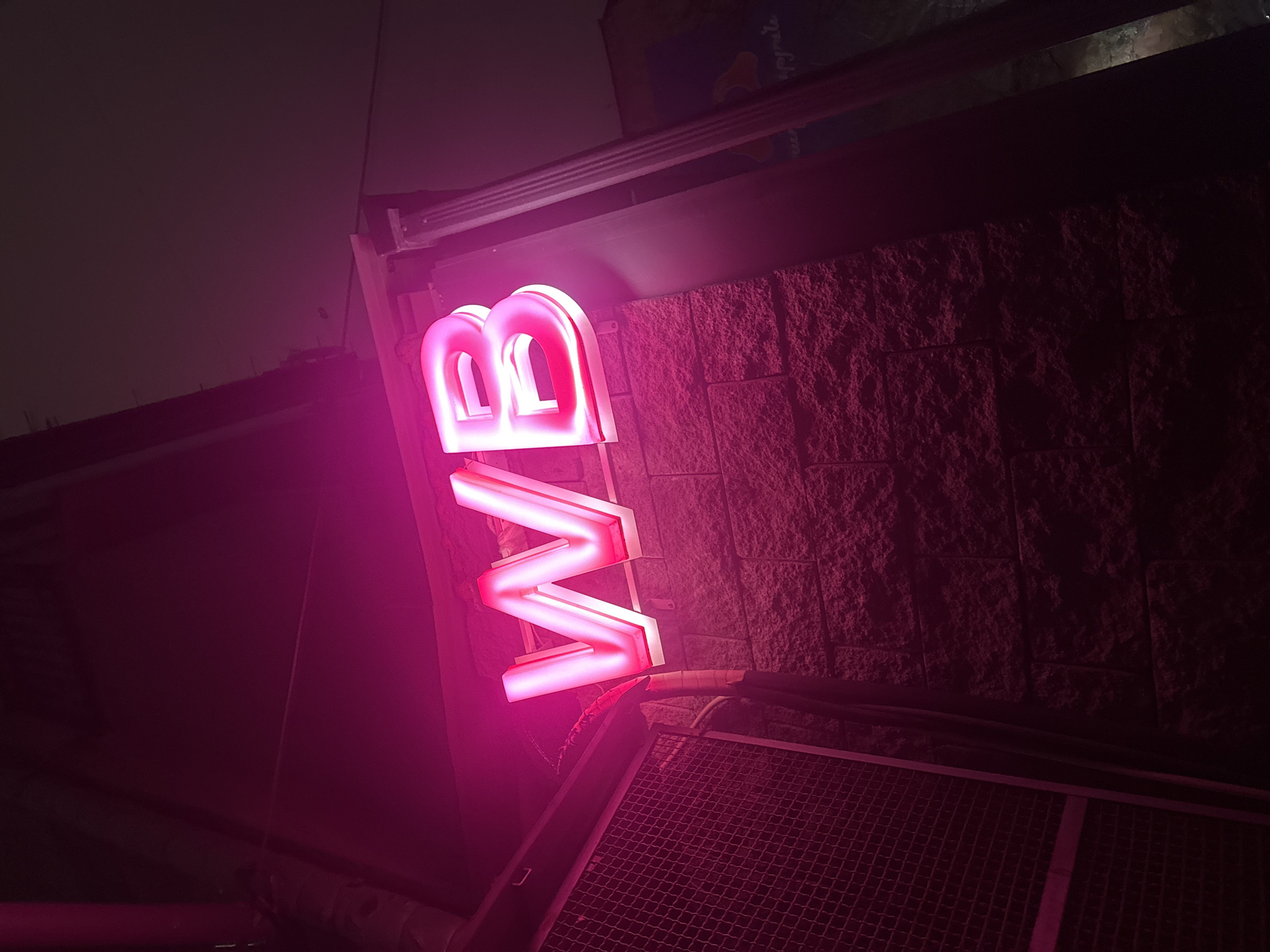 The image depicts a vertical, illuminated neon sign displaying the letters "VB" in pink. The sign is mounted on a dark, textured brick wall underneath an overhang. A metal grate is visible below the sign. The location appears to be an urban environment at night.