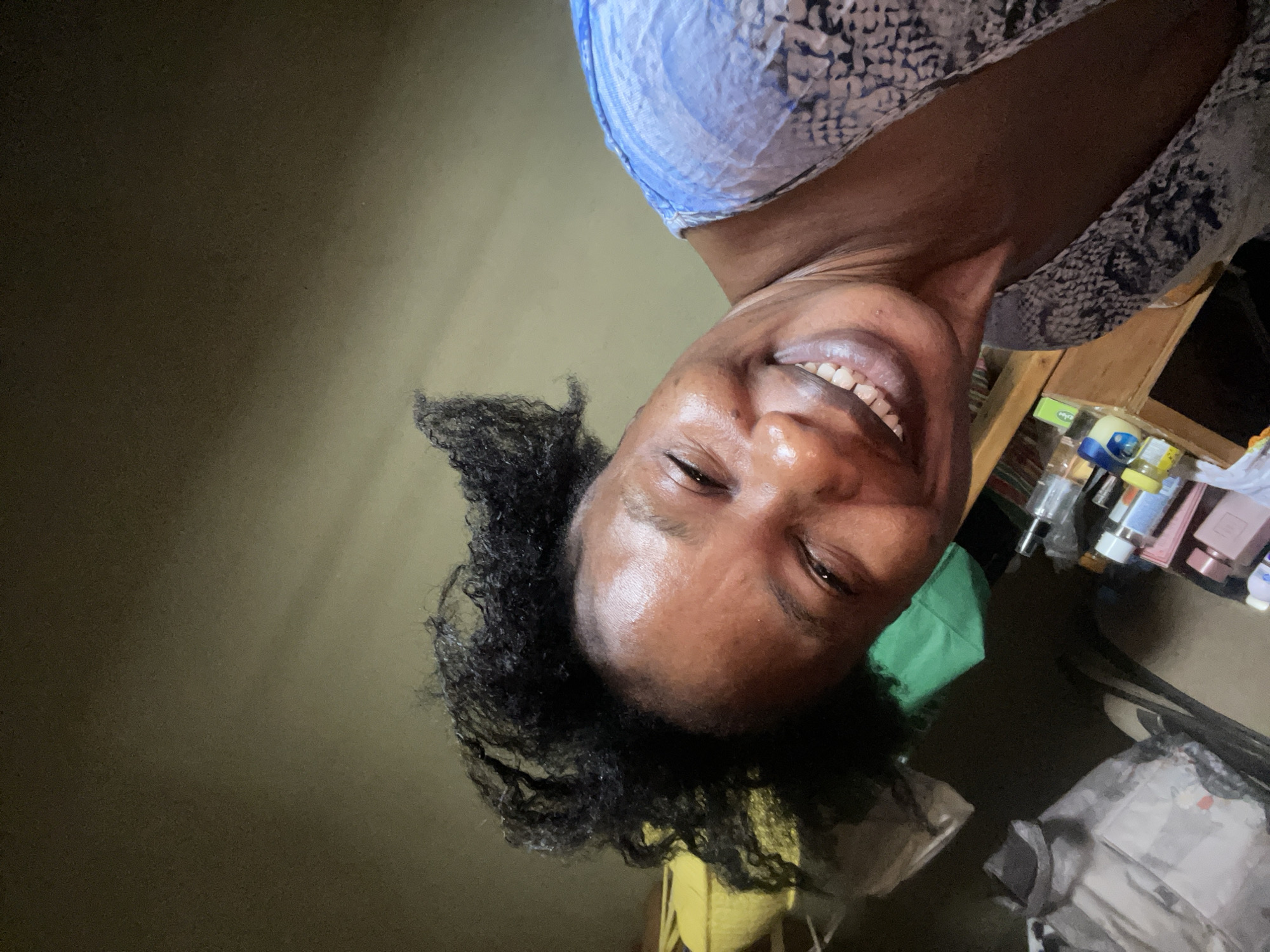 The image features a close-up, upward-angled selfie of a woman with dark skin and short, curly black hair. She is seen from the chest up, looking directly at the camera with a wide, joyful smile that shows her white teeth and causes her eyes to squint slightly. She is wearing a top with a blue and white or gray and white patterned design, possibly a snakeskin or abstract motif.
The background is split between a plain, dark olive-green wall to her right and a cluttered surface to her left. This surface appears to be a light brown wooden shelf or table laden with various personal care products, bottles, and containers. Among these items, a distinctive small white jar with a blue lid, identifiable as a "Vaseline" brand product (likely "Intensive Care"), is clearly visible. The setting suggests an indoor environment, possibly a home in Narayi, Nigeria, with warm, somewhat dim lighting that highlights the woman's face. The overall mood conveyed by her expression is one of happiness and cheerfulness.