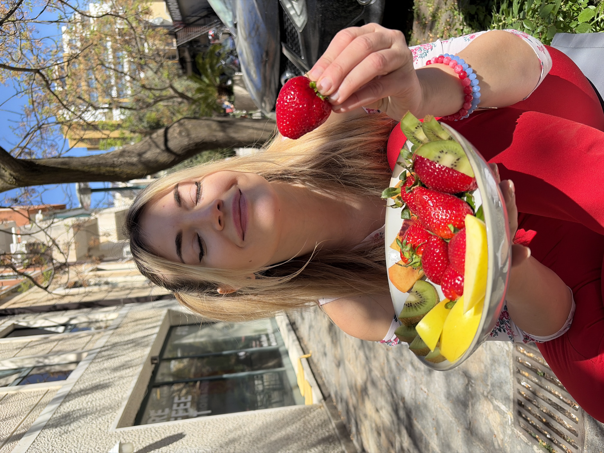 A blonde woman sits outdoors, holding a silver plate of cut strawberries, kiwi, and mango. She is also holding a strawberry in her fingers above the plate. She wears a white shirt with a floral pattern and red leggings. On her wrist, she wears two beaded bracelets in red and blue. Behind her, a building and a tree are visible. A grey car is partially visible in the background. The woman is sitting on a patterned stone surface that appears to be a sidewalk. Based on visual cues (architectural styles, foliage, surface material), the location might be in a European urban area such as Budva, Montenegro.