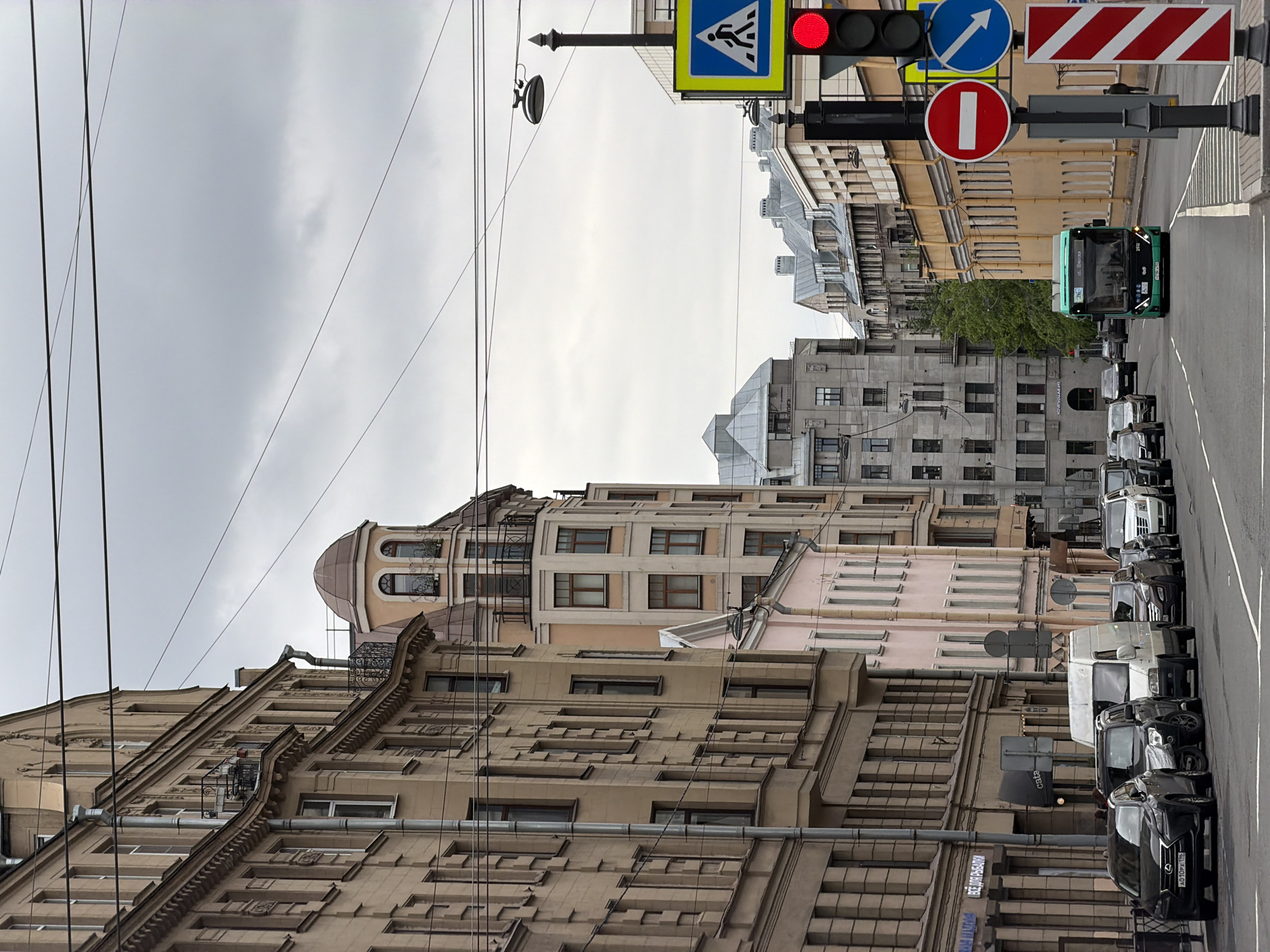 The image depicts a street scene in Saint Petersburg, Russia, characterized by a mix of architectural styles and urban infrastructure. In the foreground, the facades of multi-story buildings display elaborate details and varying colors, ranging from beige to light pink. The buildings feature numerous windows and decorative elements.
Overhead, a network of electrical wires stretches across the sky, partially obscuring the overcast sky. Street signs, including a pedestrian crossing sign and a "no entry" sign, are visible on a street lamp post.
A line of parked cars and a green bus are parked along the curb on the right side of the street. The vehicles are mostly dark-colored cars and vans.