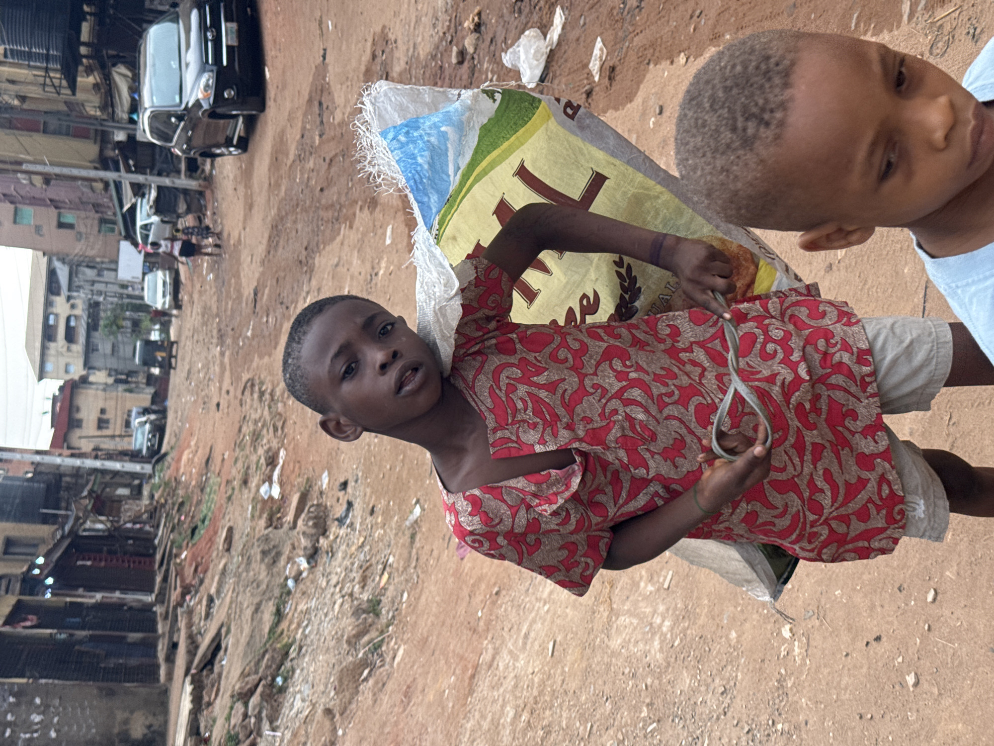 A vertical, medium-angle shot reveals a young boy, likely around 8-10 years old, carrying a large sack on his shoulder. He wears a red patterned shirt and light-colored shorts. His gaze is directed towards the camera. Another child is partially visible in the upper right corner.
The boy stands on a dirt road littered with debris. In the background are buildings and parked cars, indicative of an urban setting. The location is identified as the city of Onitsha, Nigeria. The overall scene appears impoverished.