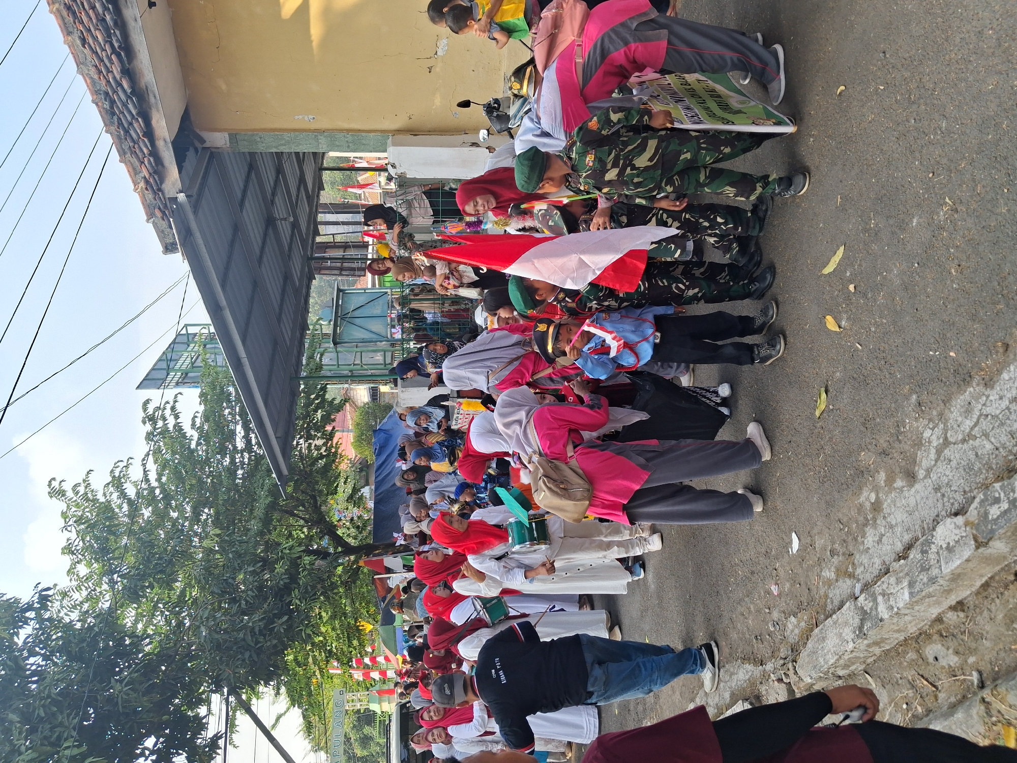 The image depicts a street scene in Balapulang, Indonesia, likely during a celebratory event or parade. A group of people, predominantly women wearing hijabs and white attire, are gathered. Some are holding green drums. Several children are also present, some dressed in military-style uniforms, complete with hats and camouflage outfits, and one child in a blue scout-like uniform. These children are holding Indonesian flags and a banner with text, the majority of which is illegible but the words "BALAPULANG" and "INDONESIA" are partially visible. The background shows a building with a yellow wall and a tiled roof, and a covered walkway. Trees and power lines are visible in the upper portion of the image. The ground is paved, with some debris scattered on it. The overall atmosphere appears to be one of community gathering and national pride.