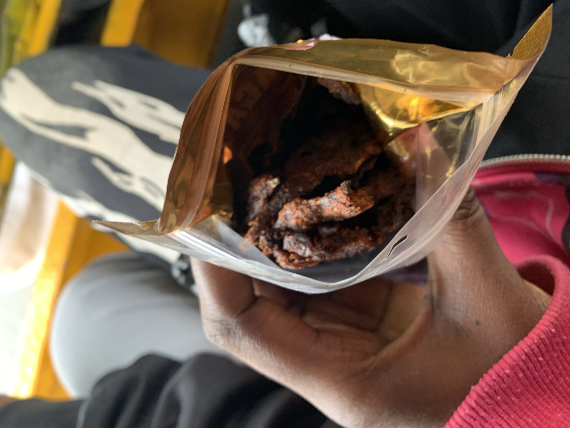 The image provides a close-up view of a person holding an open snack bag filled with dark, reddish-brown food items. The main subject is a hand, with dark skin tone, gripping a clear plastic pouch that features a prominent golden or metallic-colored interior lining. Inside the bag are numerous irregularly shaped pieces of what appears to be dried meat, possibly jerky, coated with visible spices or seasonings.
In the blurred background, partial views of clothing and surroundings are discernible. On the left, a section of dark fabric with a large, wavy white design is visible, likely part of trousers worn by the person or someone nearby. To the right, a textured red fabric, possibly a sleeve or part of a garment, is seen near the hand. The general setting suggests an indoor environment or the interior of a vehicle, with blurry yellow and dark tones in the background hinting at furniture or fixtures.
The activity shown is the holding of an open food package, implying the person is either about to eat or offering the snack. The lighting appears sufficient, suggesting daytime or a well-lit interior. No specific time of day, weather conditions, or emotional states are overtly conveyed. No discernible text is visible on the food packaging. This scene is set in Rafin Par, Nigeria.