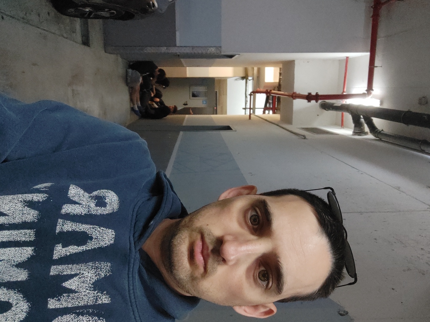 A man is taking a selfie in what appears to be an unfinished or utilitarian space. He's wearing a blue hooded sweatshirt with white lettering. Sunglasses are perched on his head.
In the background, a group of people are gathered down a long corridor. There are pipes and red fire sprinkler systems running along the ceiling and walls. The floors and walls are mostly concrete, and some sections are painted with a blue stripe. Light comes in from a distant window or opening at the end of the hall.
It is not possible to determine the location in the city of Tel-Aviv, Israel, from the image provided.
