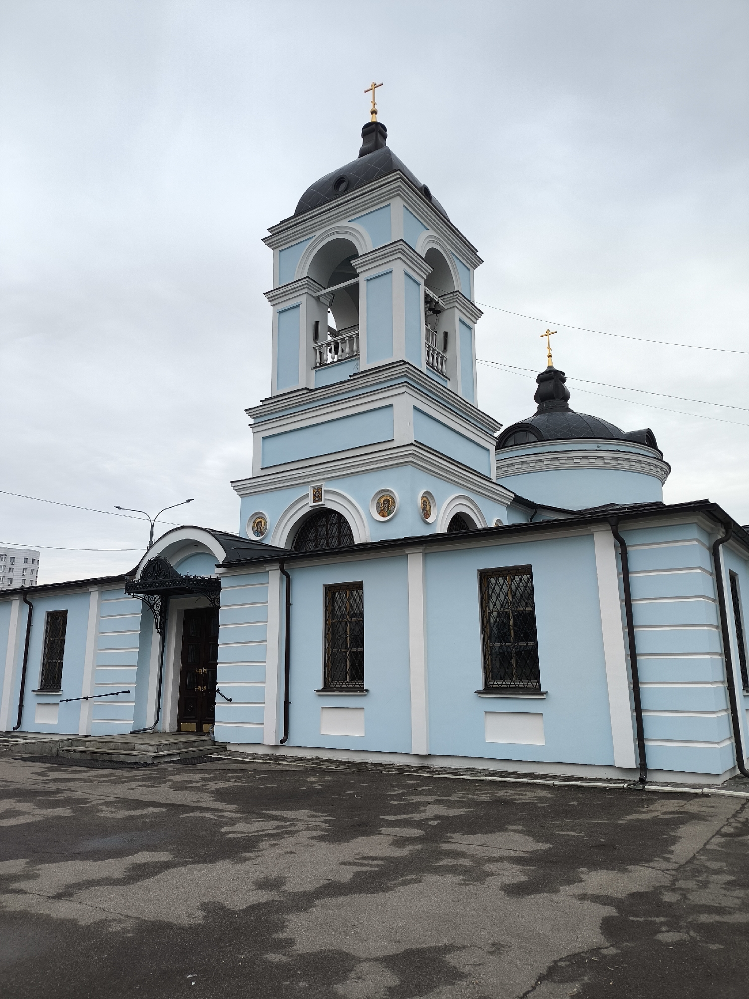 The image shows the Church of Saints Cosmas and Damian in Khimki, Russia. The church is a light blue building with white trim. It has a bell tower and a dome, both topped with gold crosses. The windows have decorative grilles. The ground in front of the church is wet asphalt. The sky is overcast.