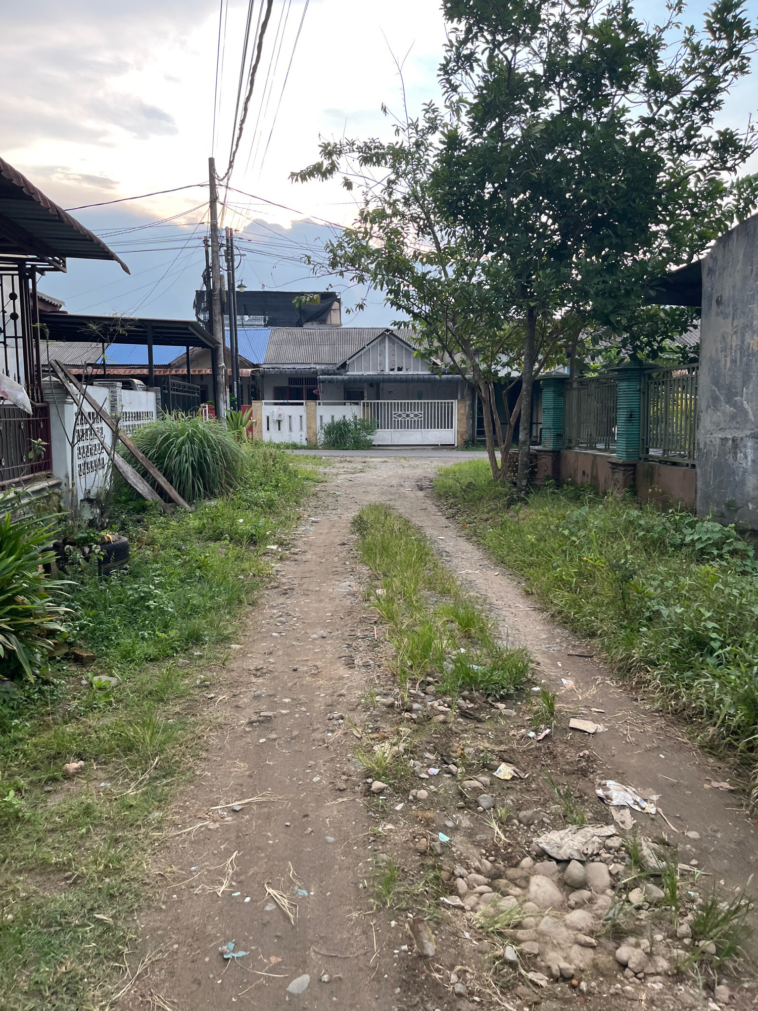A dirt path winds through a residential area in Patumbakkampung, Indonesia. The path is flanked by overgrown grass and weeds, with scattered rocks and debris, including plastic wrappers and discarded paper. On the left side of the path, a fence is visible, with a large clump of tall grass in front of it. Further back, several houses with corrugated roofs and white gates line the street. Utility poles with numerous wires are prominent in the background, indicating the presence of electricity and telecommunication services. A large tree with dense foliage stands on the right side of the path, casting shadows. The sky above is partly cloudy, with a soft glow suggesting either sunrise or sunset. The scene depicts a somewhat unkempt but lived-in urban environment.