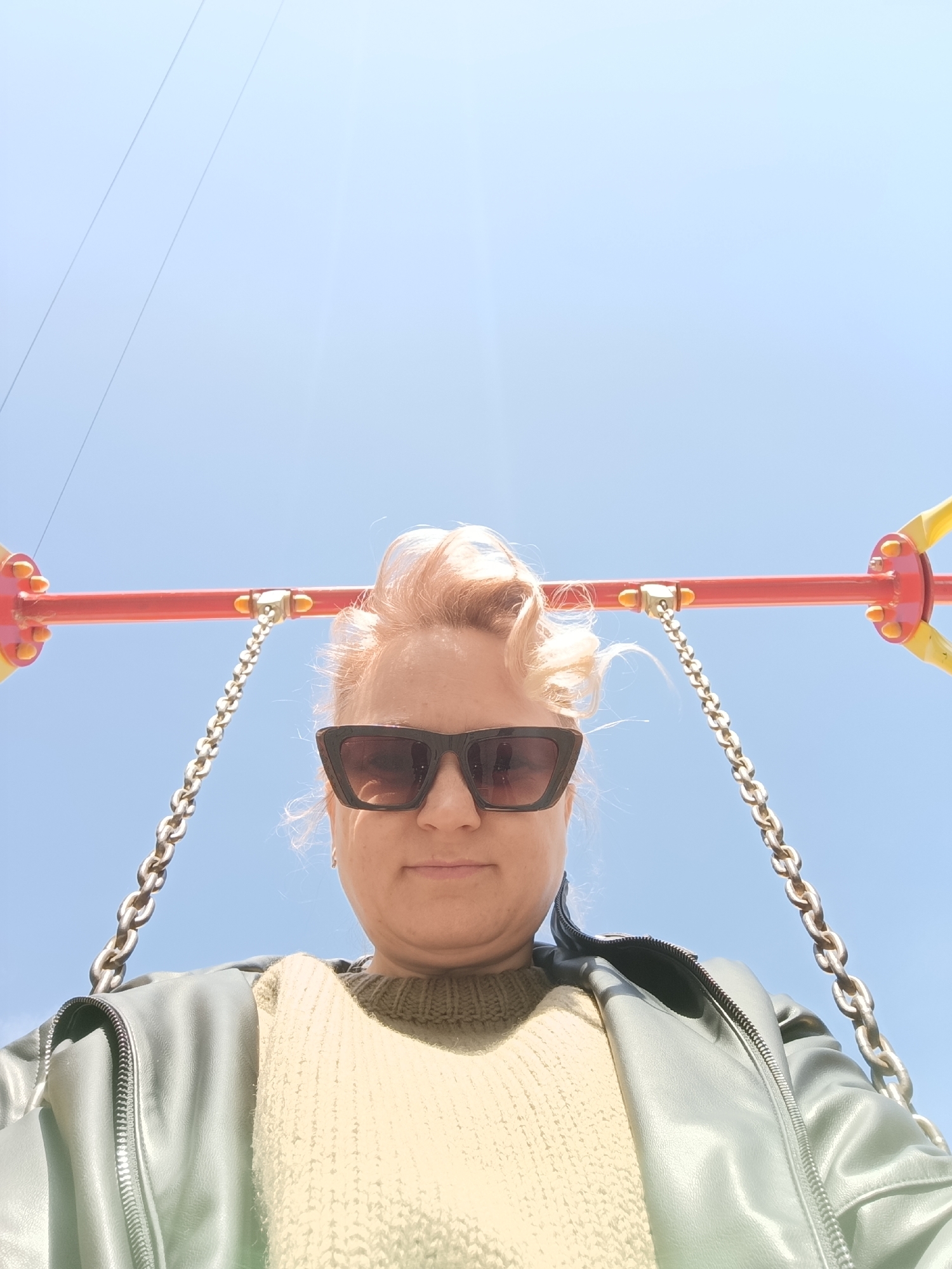 In the city of Chaikovsky, Russia, the scene depicts an individual, a woman, viewed from a low-angle perspective, positioned centrally against a clear blue sky. Her face is largely obscured by dark, rectangular-framed sunglasses. She has light blonde hair, styled with some volume. She is wearing a light olive-green knitted sweater beneath an open, light grey-green faux leather jacket. Directly above and behind her, a red horizontal bar, consistent with a swing set structure, spans the upper portion of the frame. This bar exhibits yellow plastic caps or decorative elements at its ends. Two metal chains, suspending from this red bar, are visible on either side of the woman's head, indicating she is seated on a swing. Faint, parallel utility lines are visible against the blue sky in the upper left quadrant. The environment is outdoors, under bright daylight conditions.