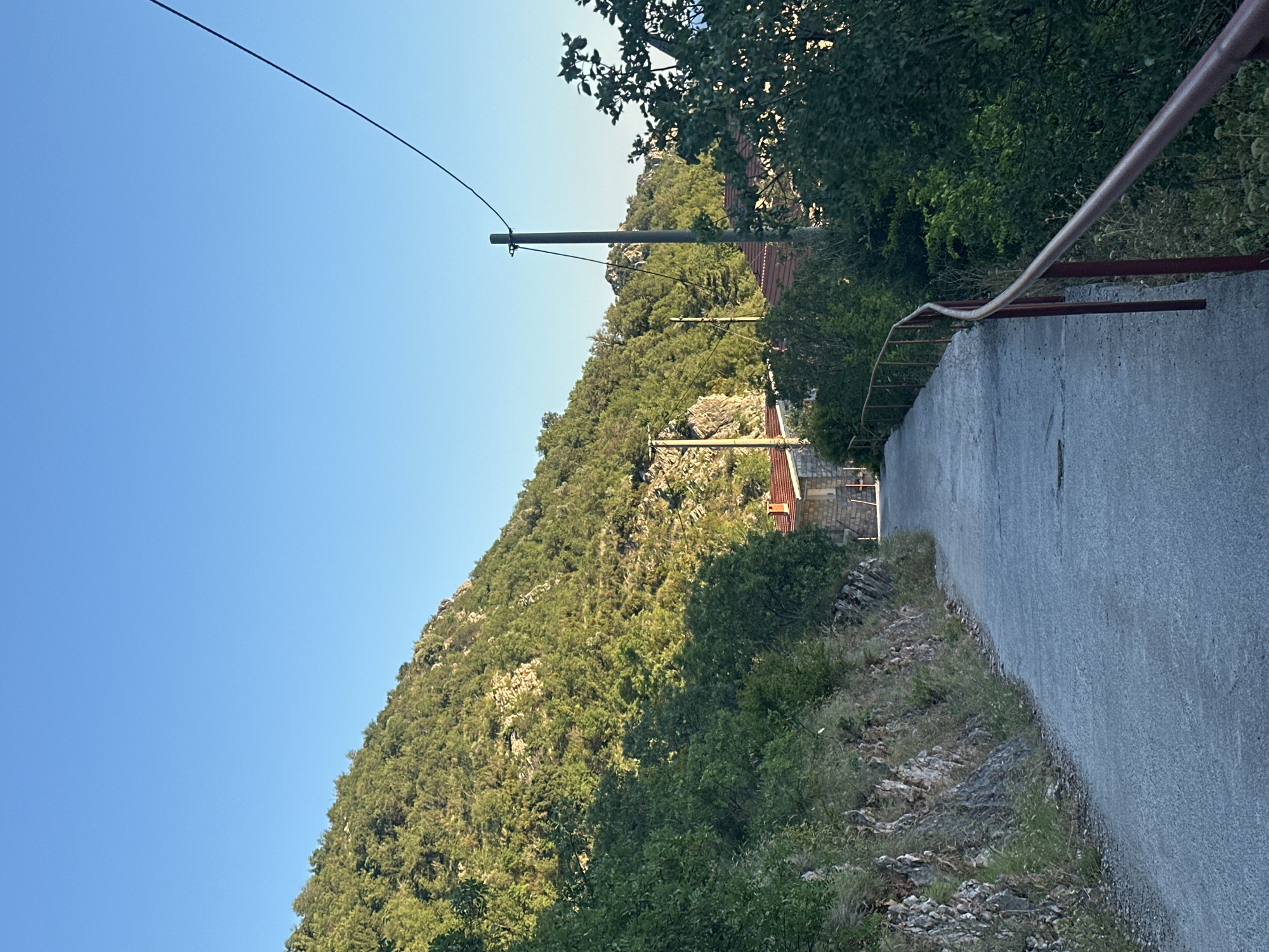 The image depicts a paved pathway winding upwards through a hillside landscape near Čučuci, Montenegro. The path is bordered on the left by a rocky embankment covered in sparse vegetation, and on the right by a red metal railing. Lush green trees and bushes line the sides of the path and cover the steep hillside that dominates the left and upper portions of the frame. In the distance, glimpses of red-tiled roofs indicate the presence of buildings nestled among the foliage. A utility pole with wires is visible against the clear blue sky. The scene appears to be captured in natural daylight, likely during the late afternoon or early morning, given the long shadows and the warm illumination of the vegetation. There are no people or animals visible in the image.