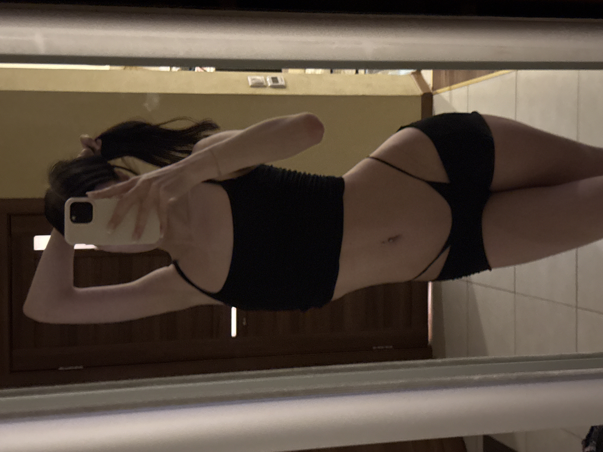 In a dimly lit indoor setting, a person is standing in front of a mirror, taking a selfie with a smartphone. The person is wearing a black crop top and black shorts. Their hair is pulled back in a ponytail. The environment appears to be a room with a door and tiled floor visible in the reflection. The location is identified as the city of Khimki, Russia.
