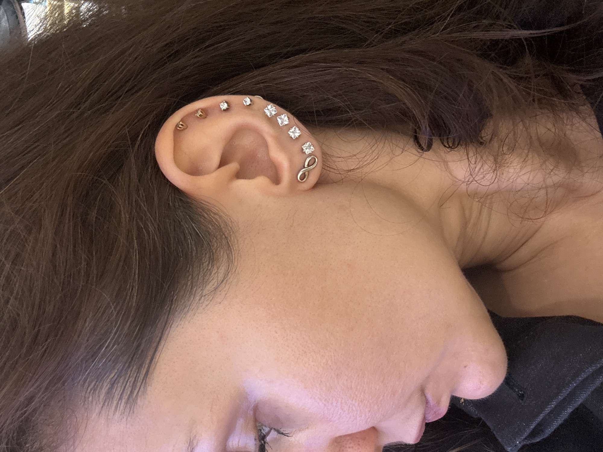 A close-up image captures the left side of a person's face. The focus is on the ear, which is adorned with several piercings. These include a row of small, round studs along the upper ear and a group of square, gemstone-like earrings near the top. At the lower edge of the ear, there is a distinctive infinity symbol earring. The person has dark brown hair that frames the face, and the skin tone is light. The person's eyes are closed, and the position of the head suggests they are lying down. The person is wearing a dark-colored garment that is visible at the neck and shoulder area. The background is softly blurred and indistinct. There is no indication of the location being Dubai, United Arab Emirates.